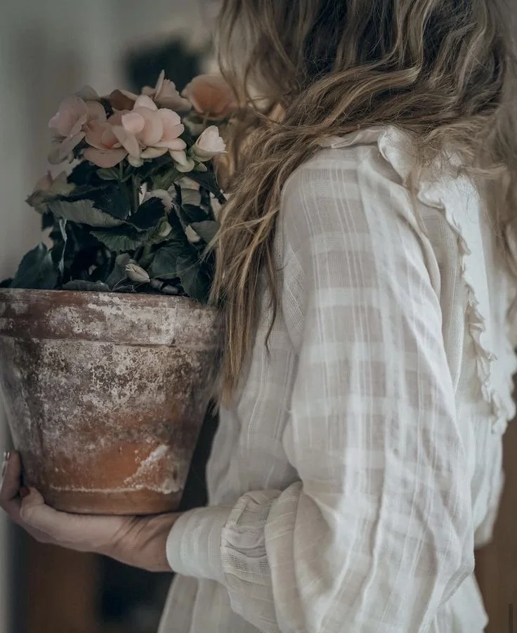 A person with long, wavy, blonde hair holding a large, weathered terracotta pot with pink artificial flowers and green leaves.