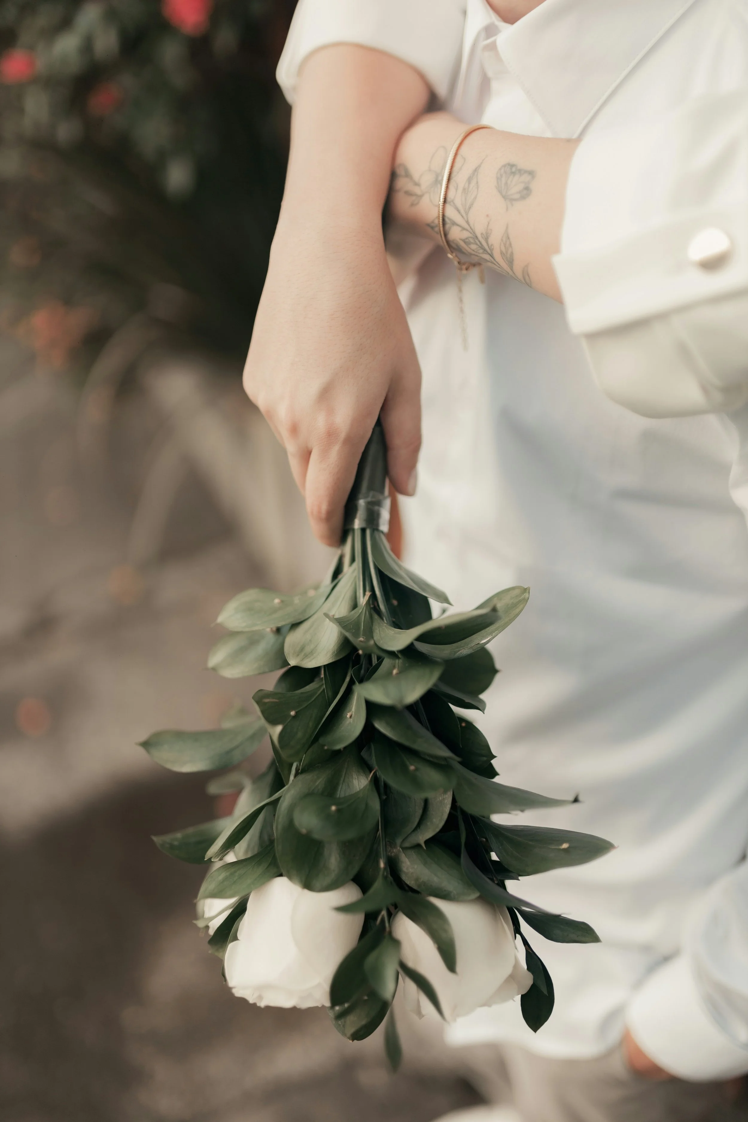 Person holding a bouquet of greenery and white flowers, wearing a white blouse with rolled sleeves and a gold bracelet, standing outdoors.