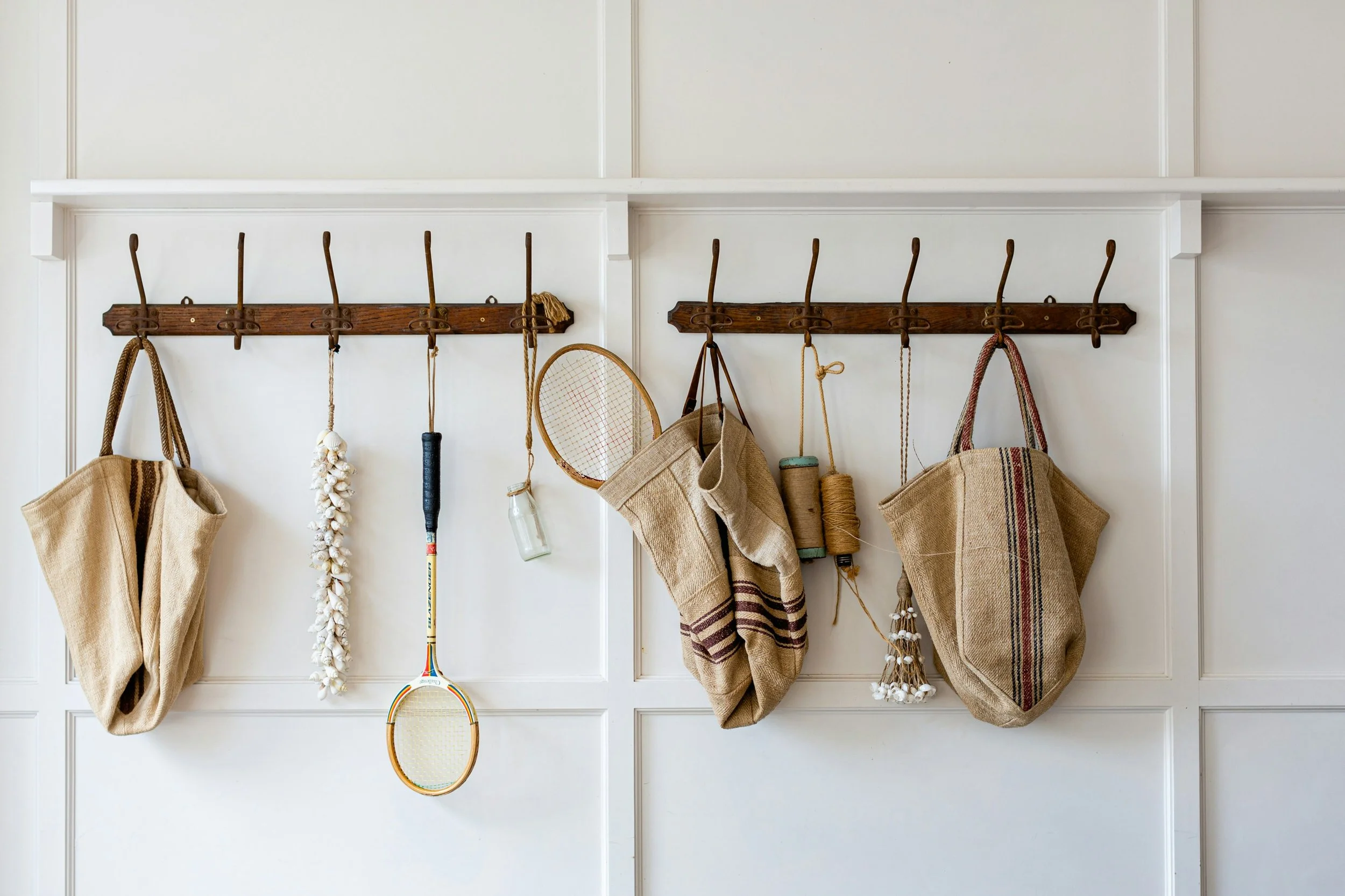 A white wall with two wooden coat racks, each with five hooks. On the hooks hang beige and striped bags, a tennis racket, a bulb, and other items.