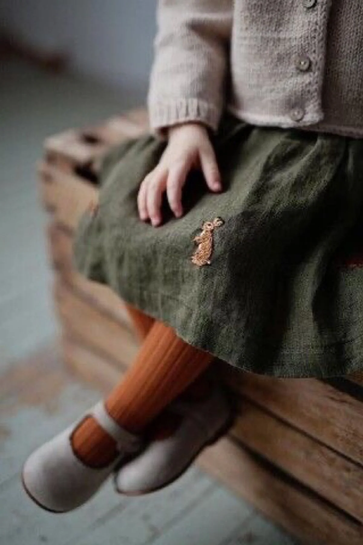 A child's hand rests on their lap, wearing a brown sweater and olive-green shorts with a small embroidered bear patch. The child is sitting on a wooden chair with a white socked foot hanging down.