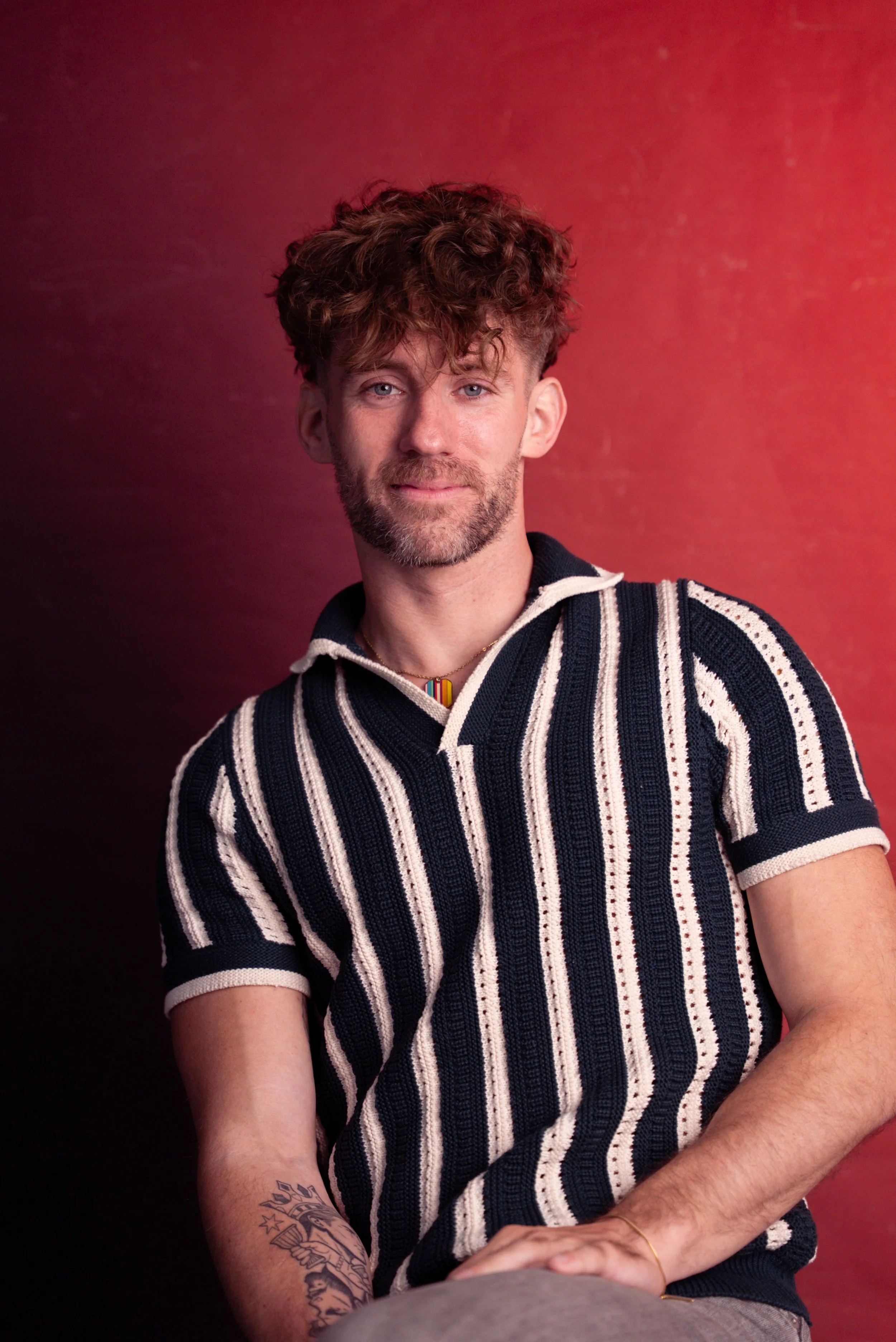 A young man with curly brown hair, beard, and light skin, wearing a striped black and white shirt, sitting against a red background.