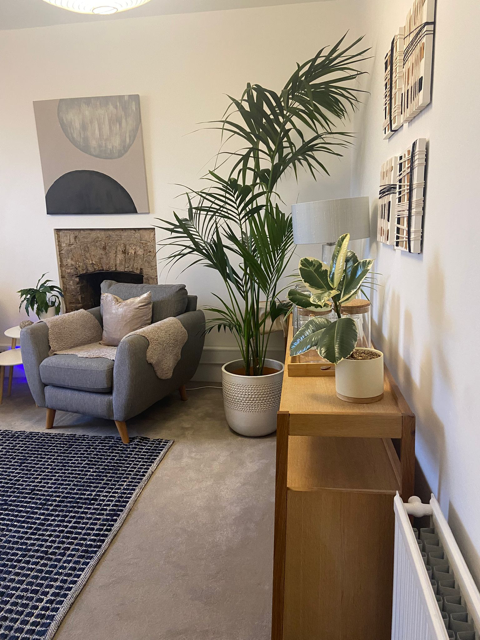 Living room corner with a gray armchair, large indoor plant, wooden console table with potted plants and a table lamp, framed wall art, and a fireplace with a decorative stone surround.