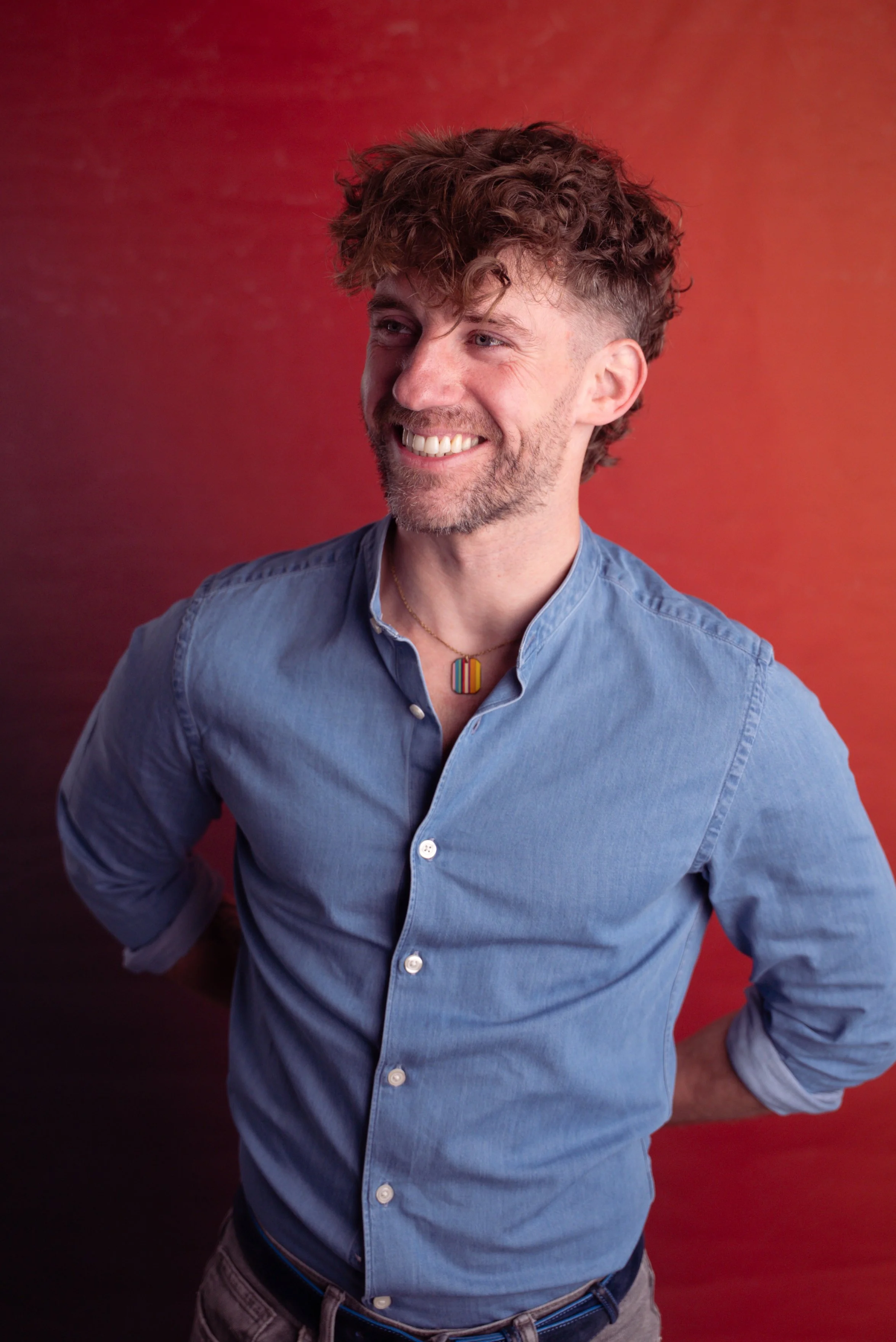 A smiling male therapist with curly brown hair, wearing a light blue button-up shirt and a colourful rainbow-striped necklace, standing against a red background.