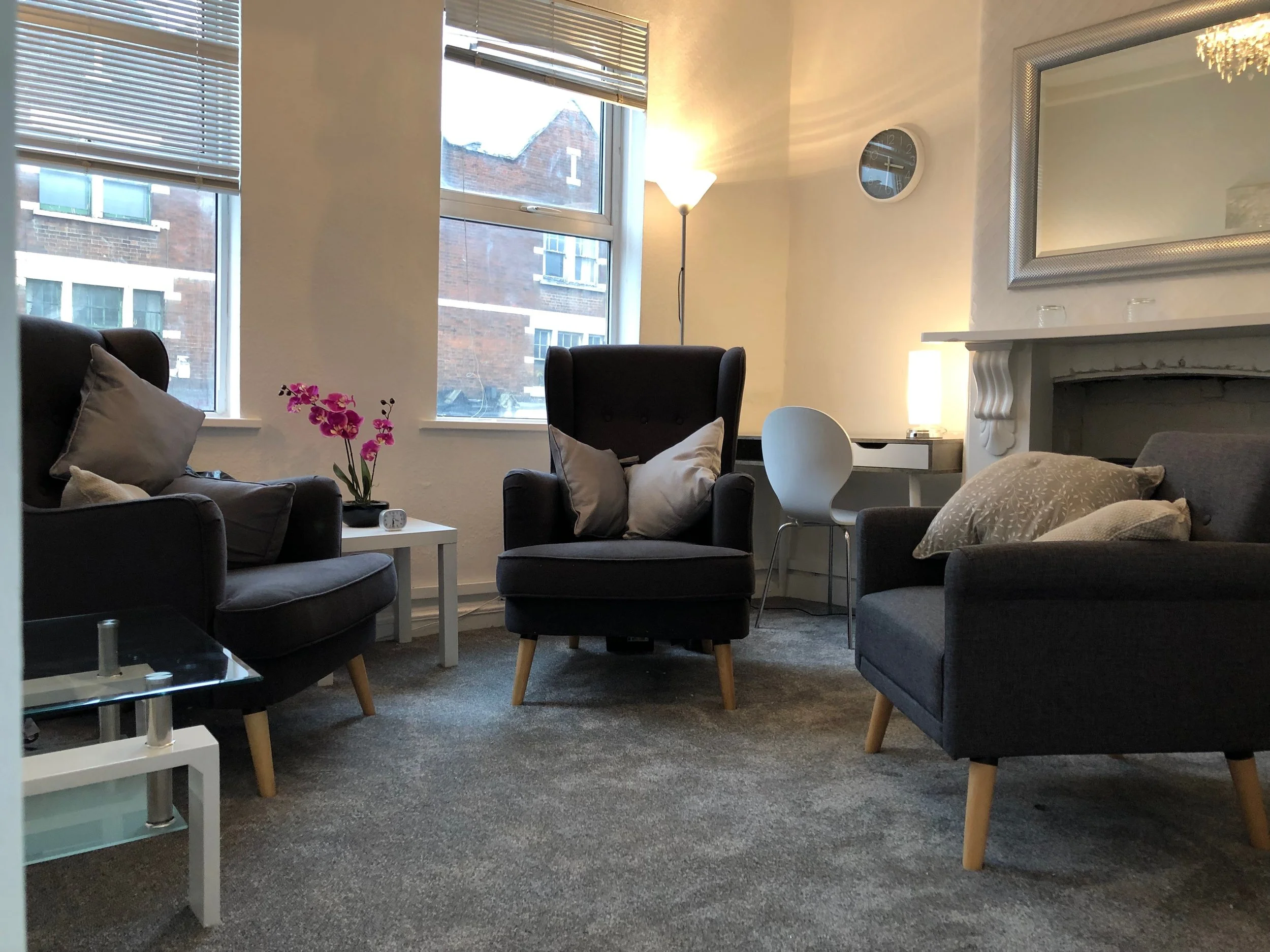 A cozy living room with two dark armchairs, pillows, a white side table with a pink orchid, a glass coffee table, a white desk with a white chair, a silver-framed mirror, a round white wall clock, a floor lamp with a white shade, and large windows with blinds showing neighboring brick buildings.