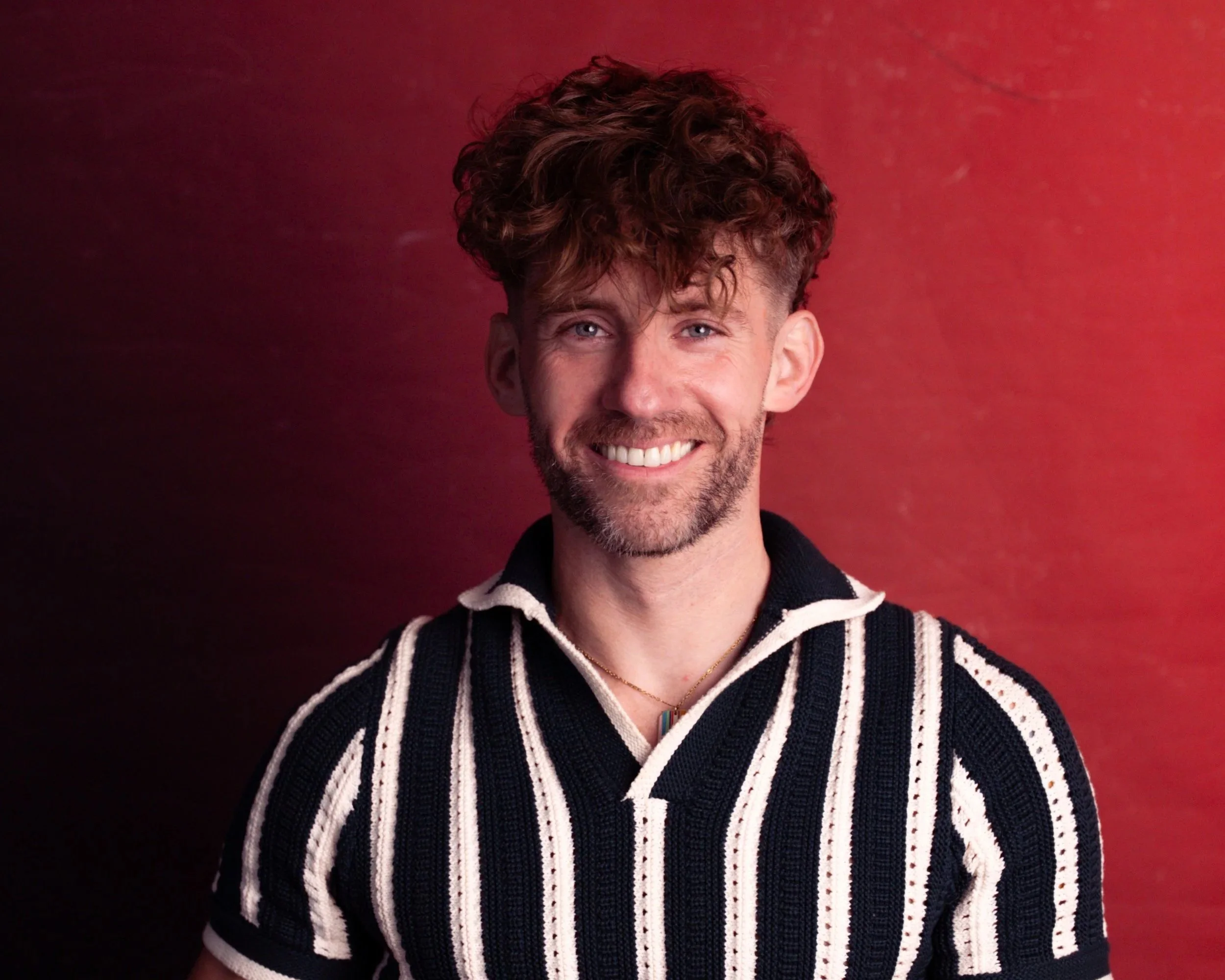 A young man with curly brown hair, light blue eyes, and a beard, smiling against a red background, wearing a black and white striped sweater.