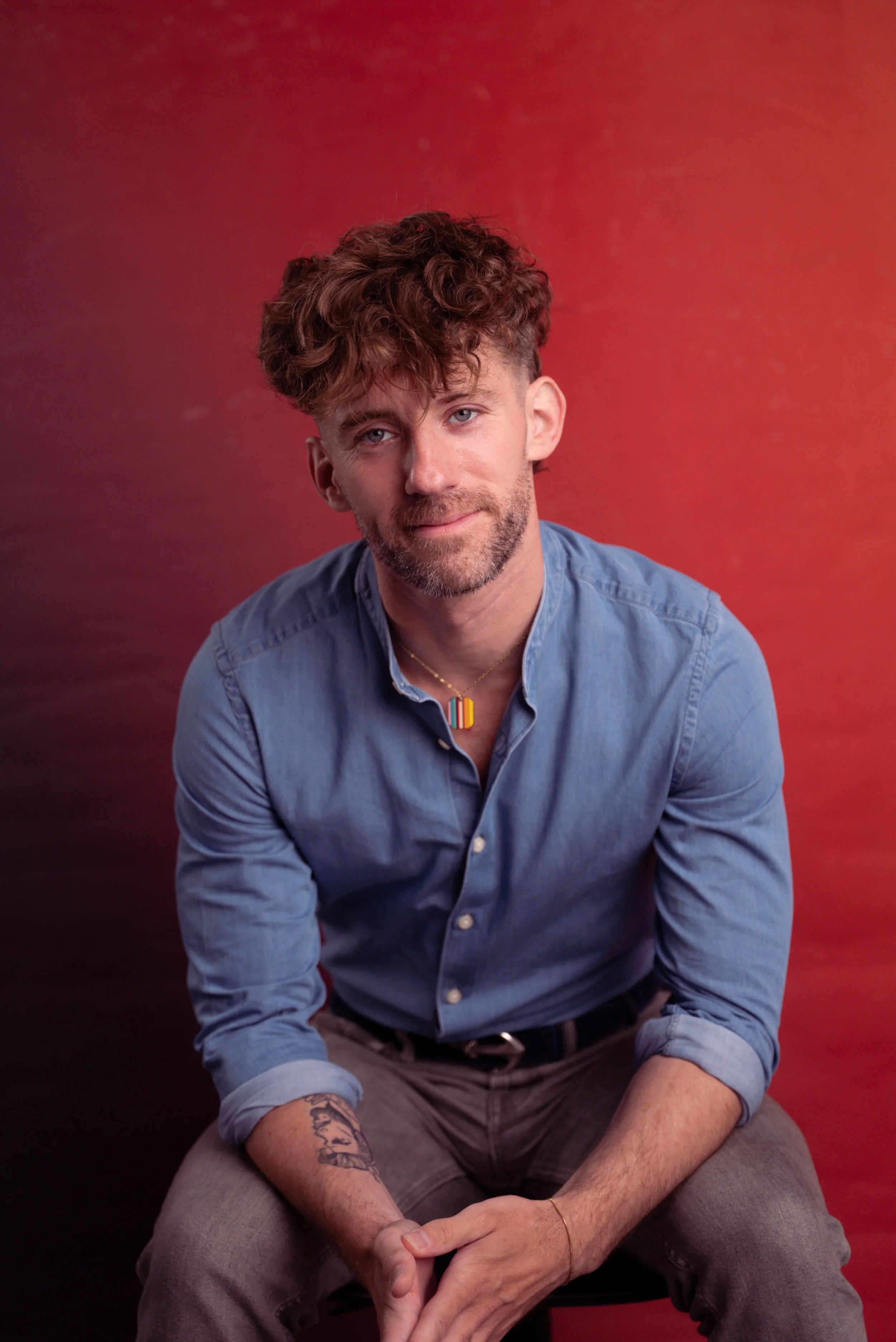 A young man wearing a light blue shirt sitting against a red background.