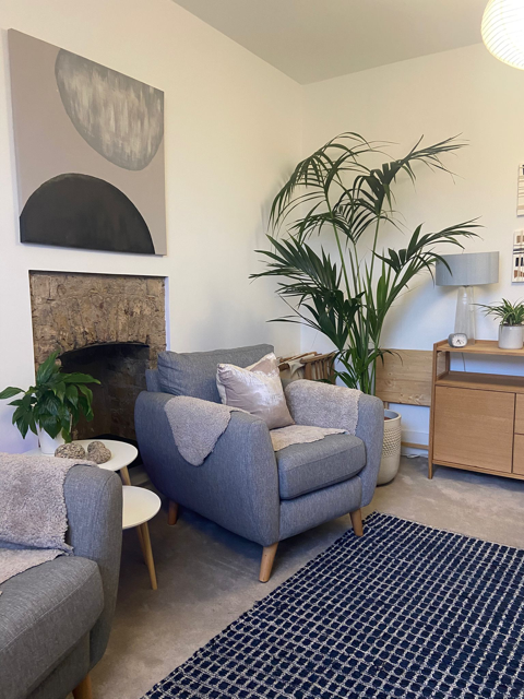Living room with gray armchair, potted plants, abstract black and white wall art, fireplace, wooden sideboard, lamp, and patterned rug.