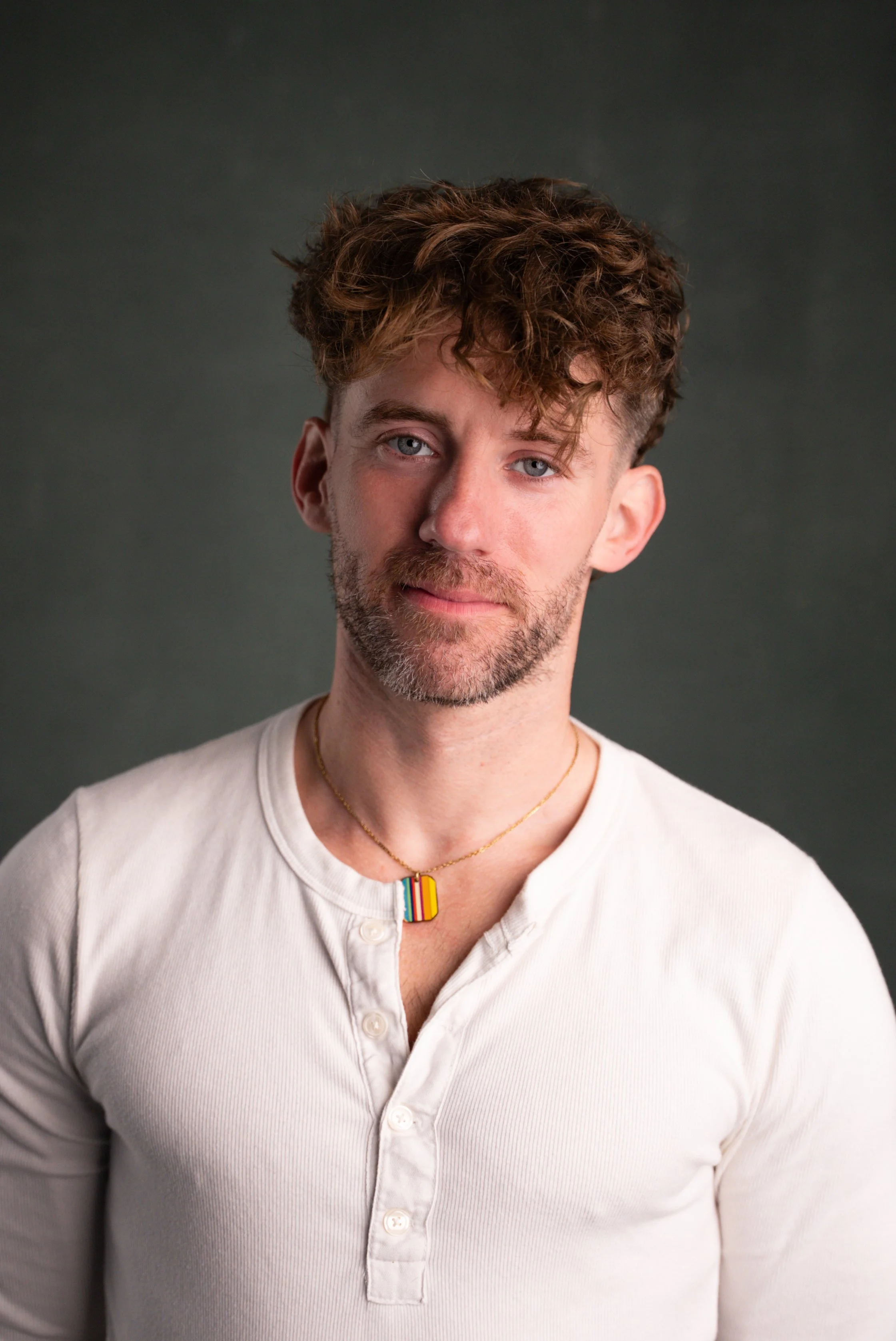 A male therapist with wavy brown hair, blue eyes, and a beard wearing a white button-up shirt and a rainbow-colored pride necklace, standing against a dark background.