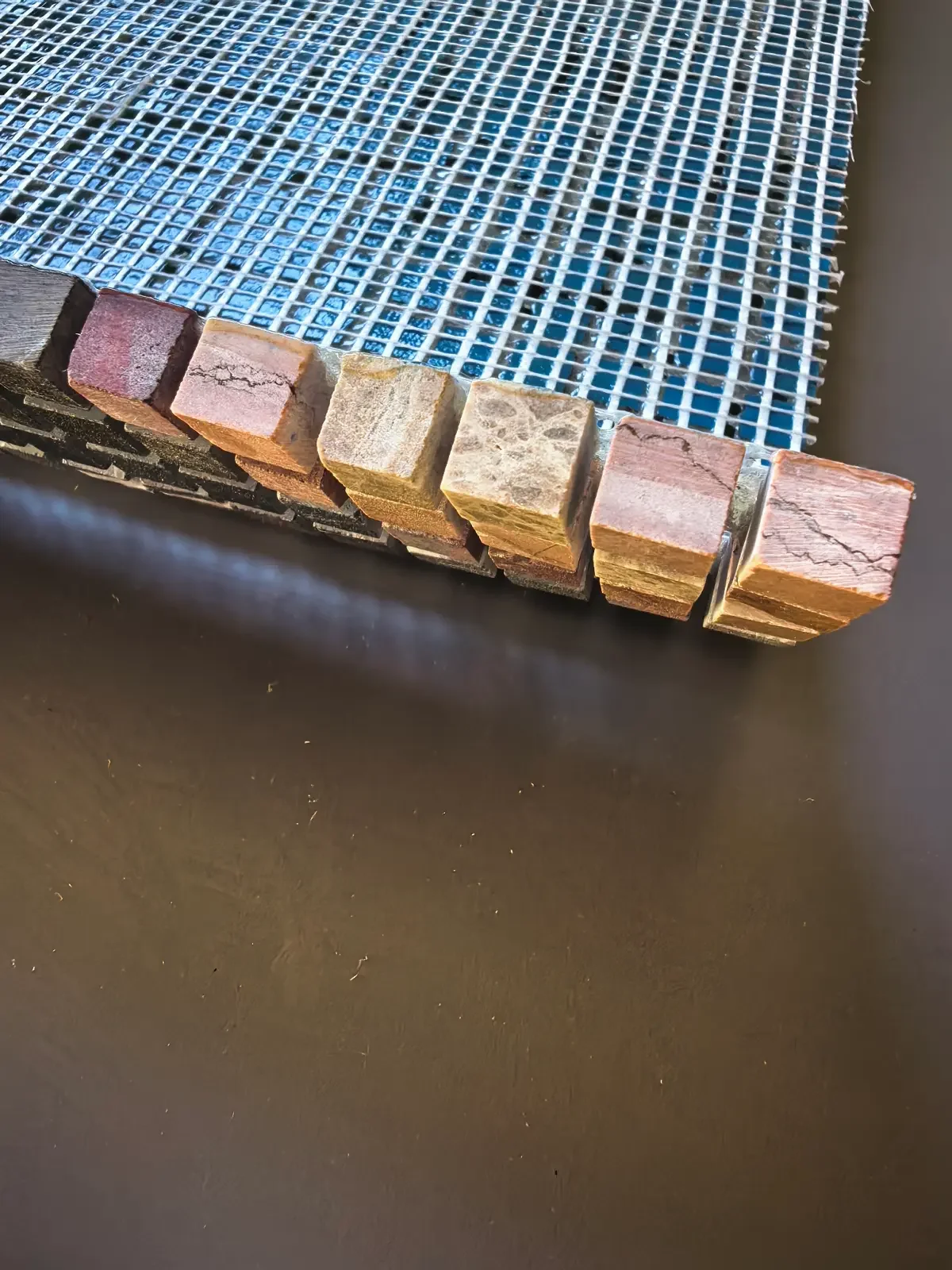 Edge of a modular mosaic panel on its mesh backing, showing stone tesserae in warm marble and travertine tones
