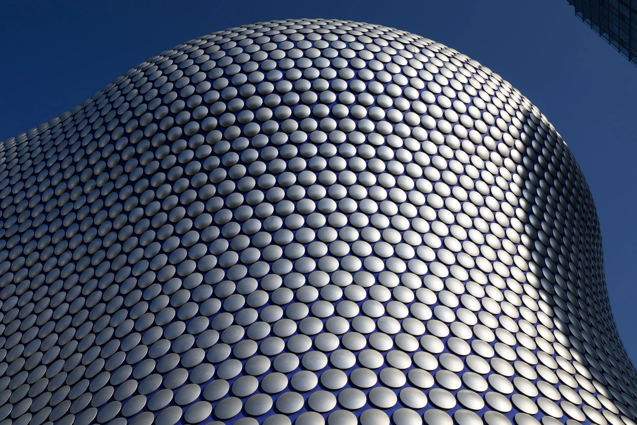 Close-up of a modern building's rounded exterior wall covered with white circular tiles, with a tall glass building visible in the background against a clear blue sky.