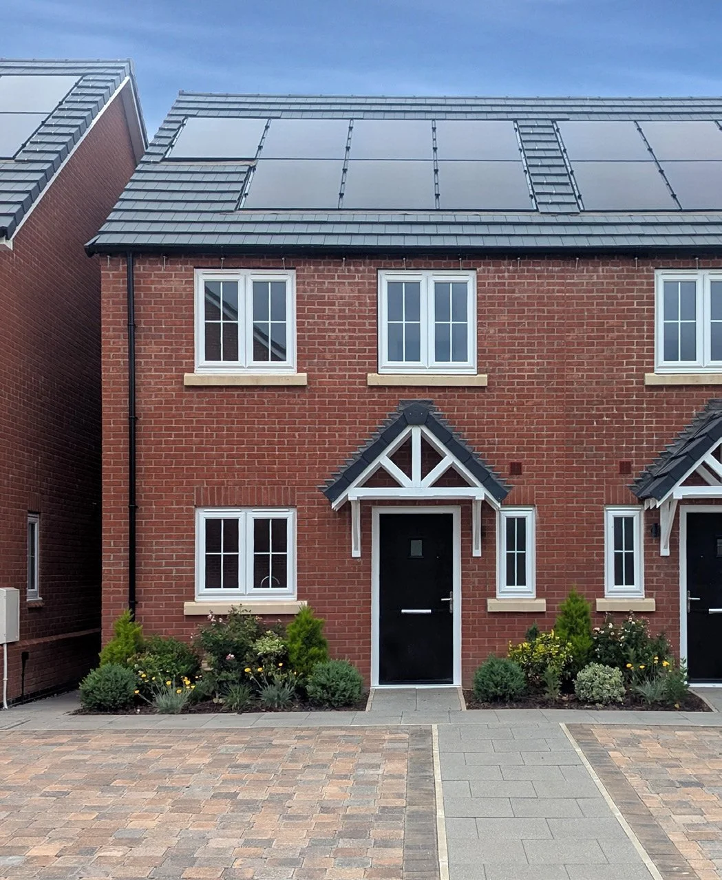 A modern brick house with solar panels on the roof, black front door, white window frames, small front garden with bushes and flowers, and a paved driveway.