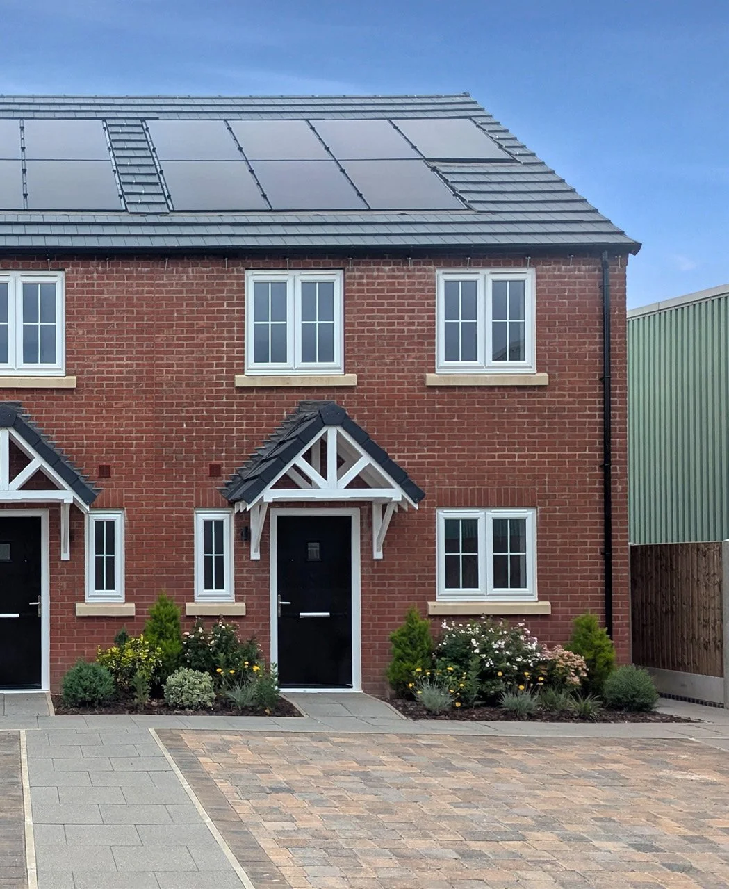 A modern brick house with solar panels on the roof, white-framed windows, and a black front door with a small porch and landscaped garden.
