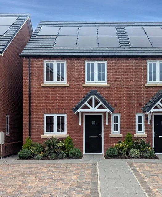 Red brick house with black roof, white window frames, and a black front door, with small garden beds and a paved driveway.