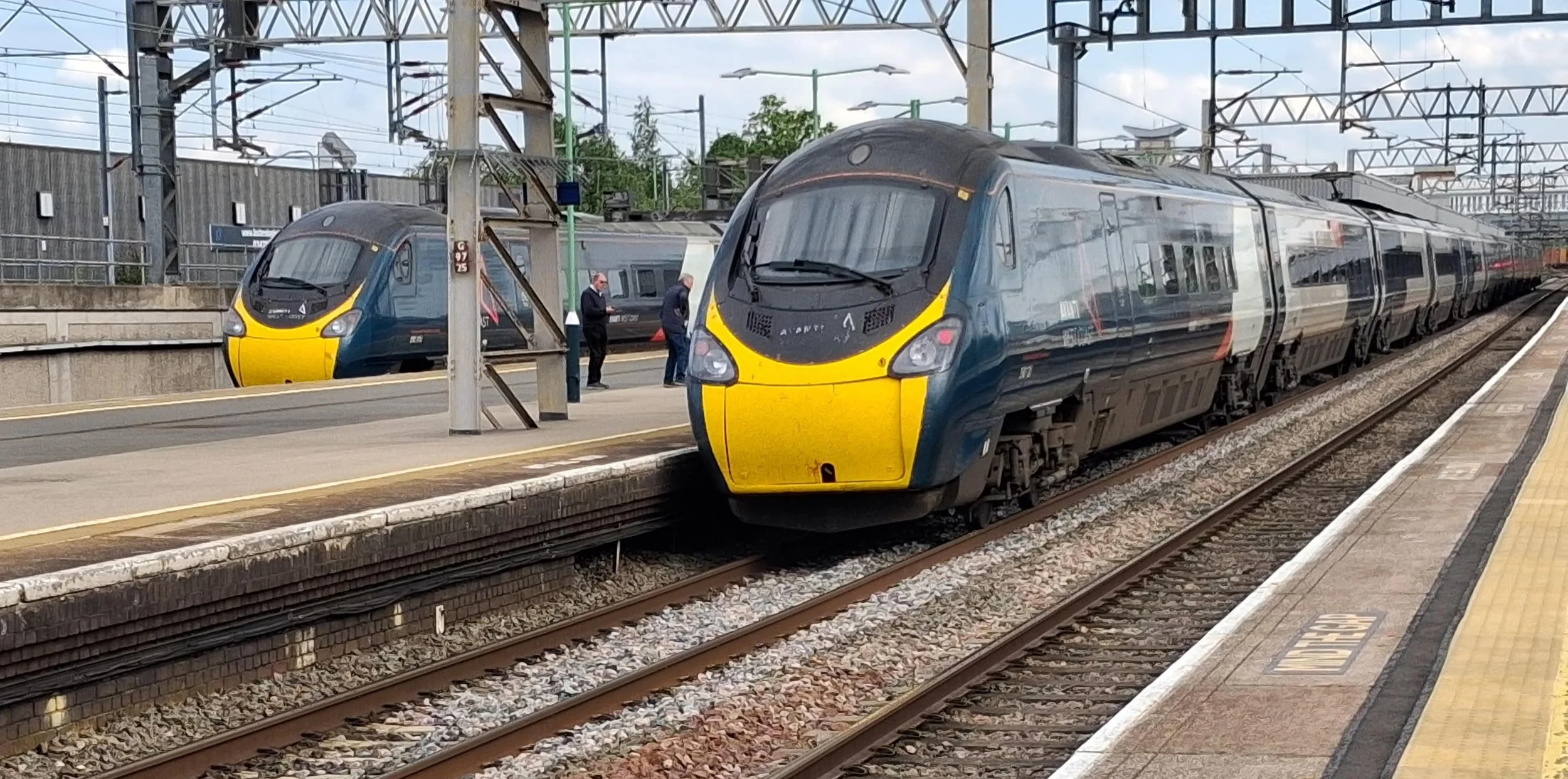 Two modern trains are at a train station platform, with one train arriving and the other standing still in the background. The trains have a sleek, curved design with blue and yellow colors. Two people are standing on the platform, engaged in conversation. The station has overhead electrical wiring and a concrete platform with yellow safety lines and tactile paving.