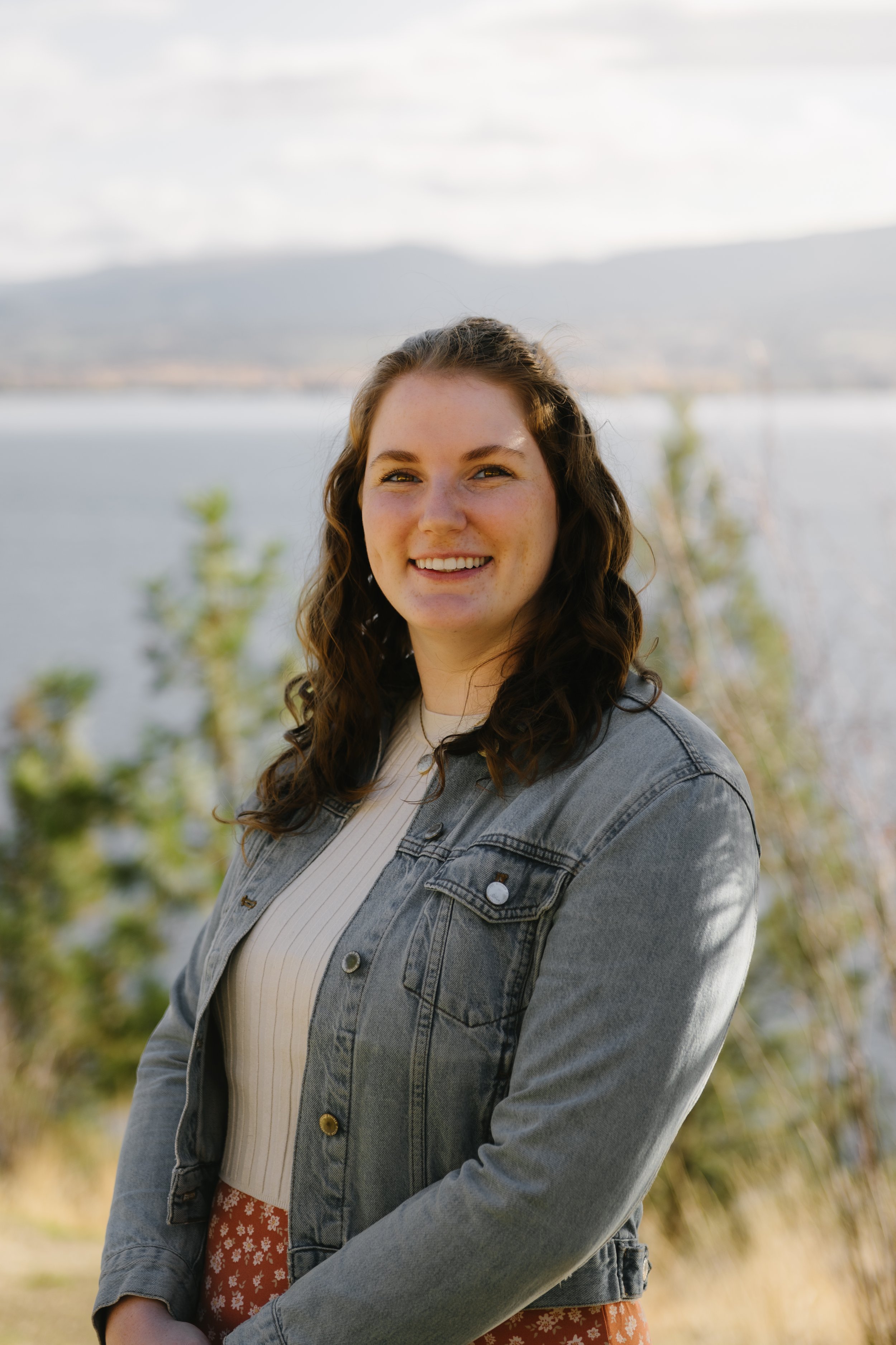 A woman with curly brown hair smiling outdoors near a body of water with mountains in the background.