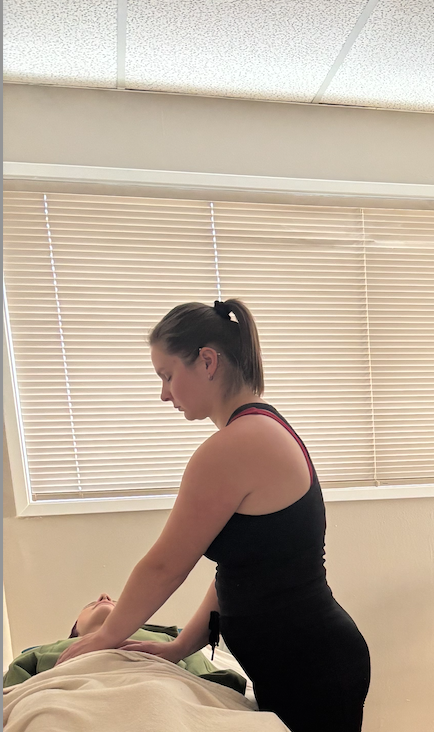 A woman giving a massage to a person lying on a bed in a room with closed blinds.