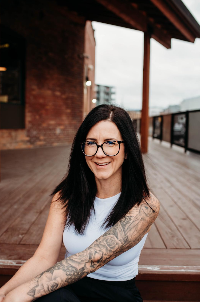 A woman with long black hair, tattoos on her arm, and glasses, smiling while sitting outdoors on a wooden deck.