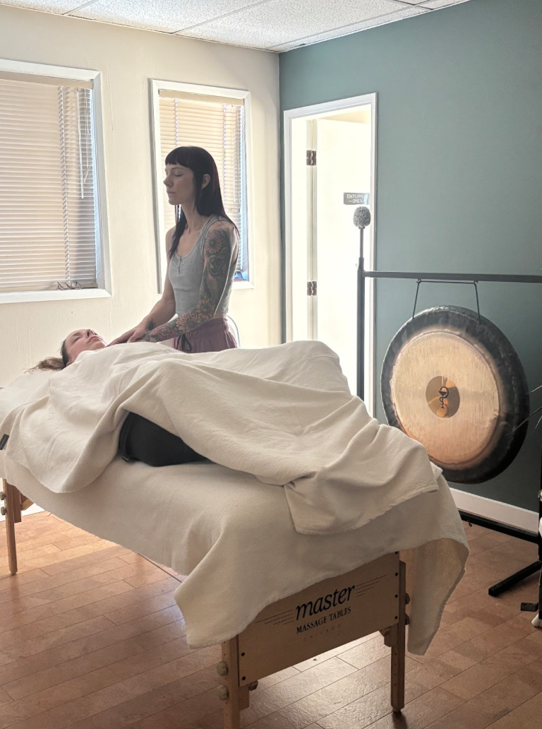 A woman with tattoos on her arms performing a massage on another woman lying on a massage table in a room with light-colored walls and windows with blinds. A large gong is visible in the background.