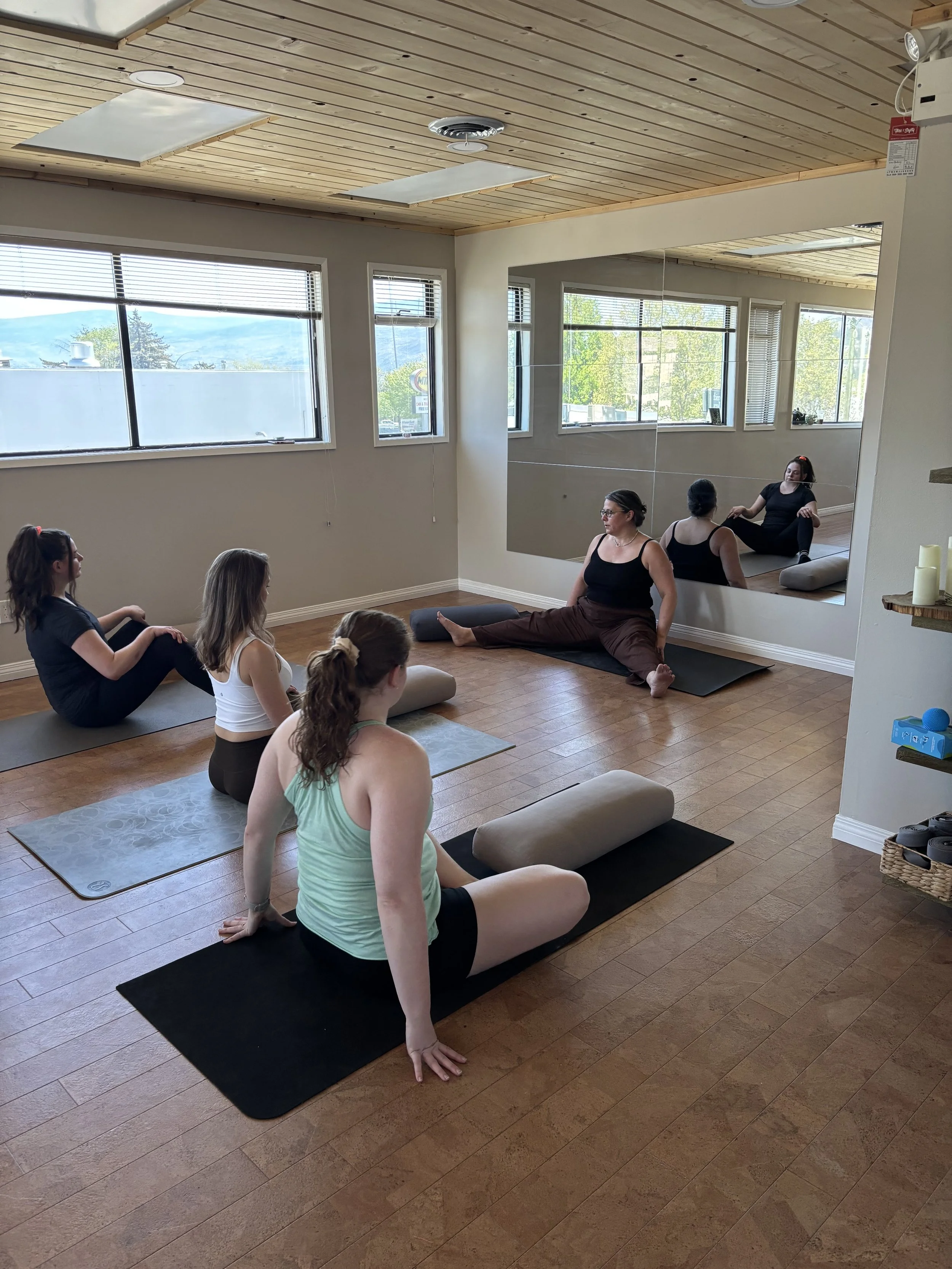 A group of women participating in a yoga class in a studio with wooden floors and a mirrored wall. They are sitting on yoga mats and cushions, some in seated poses and others in a stretch with one leg extended and the other bent, using their hands to bolster themselves. Large windows show a scenic view of trees and mountains outside.