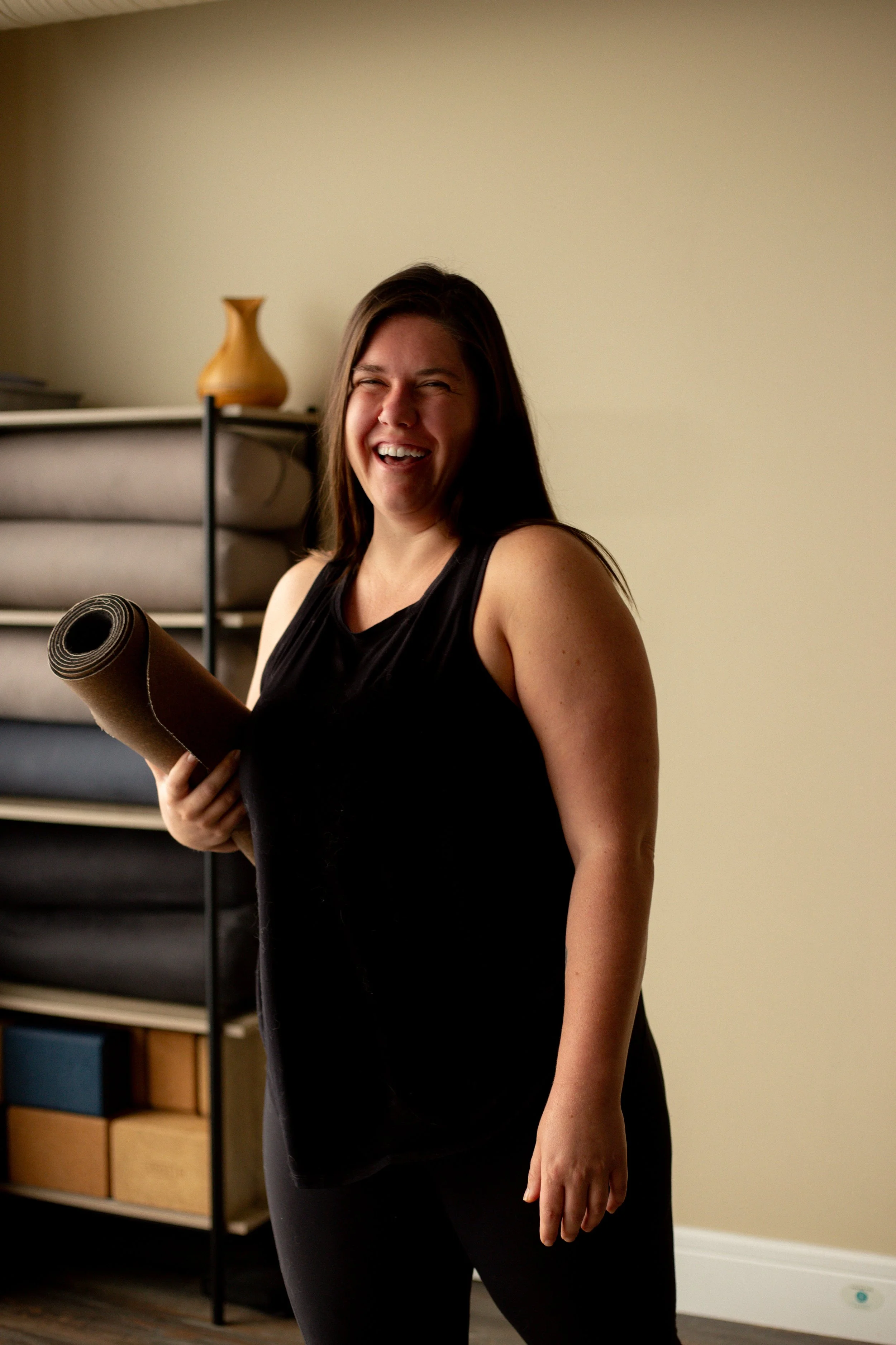 A woman with long brown hair, wearing a black sleeveless top, holding a rolled-up yoga mat, smiling and standing in a room with a beige wall, a bookshelf with various storage boxes, a shelf with a wooden vase, and a rolled-up yoga mat.