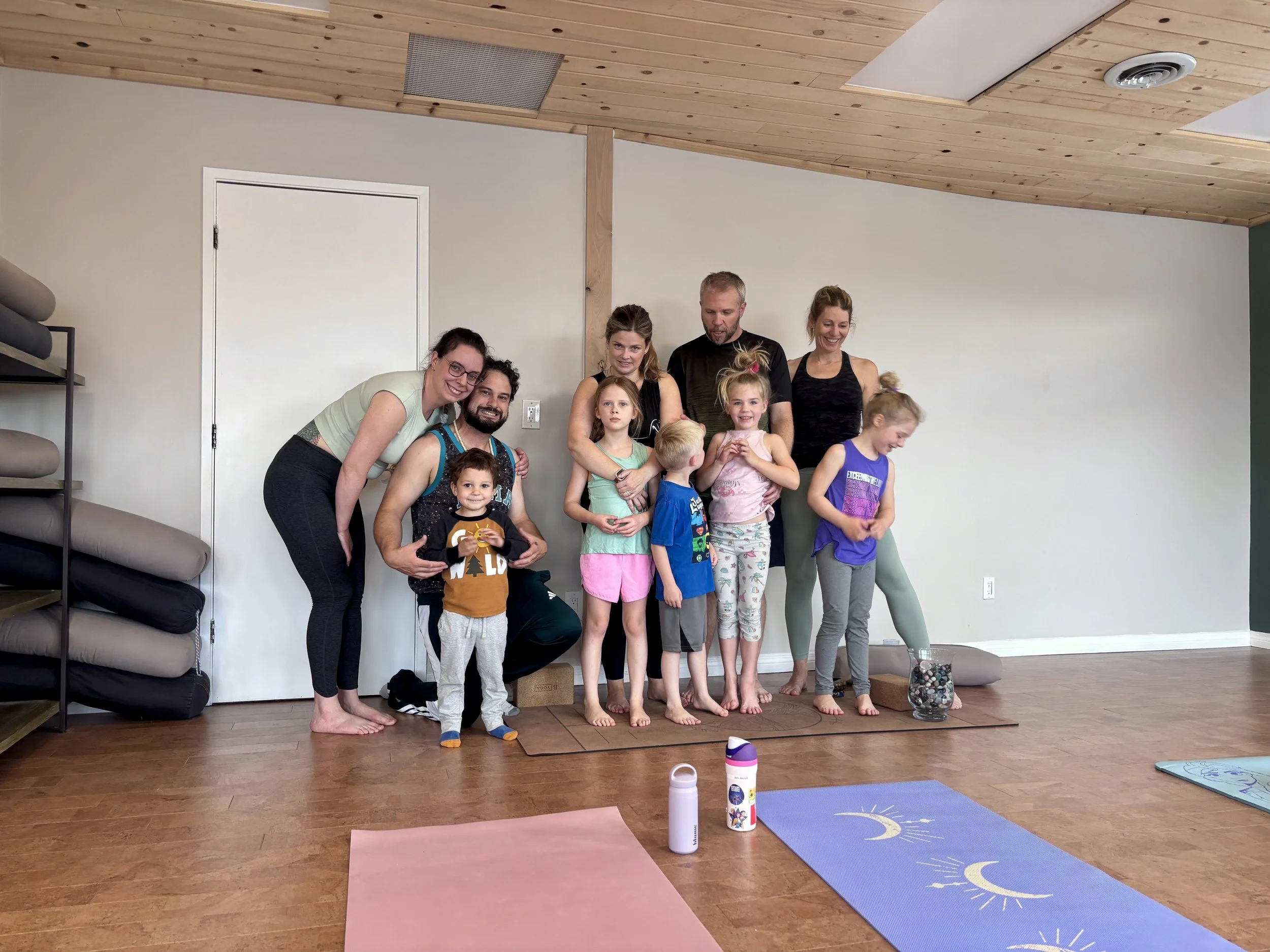 A group of people, including adults and children, gathered together indoors for a yoga or meditation class, standing on a cork board floor with yoga mats and bottles in front of them, against a plain wall with a door and shelves in the background.