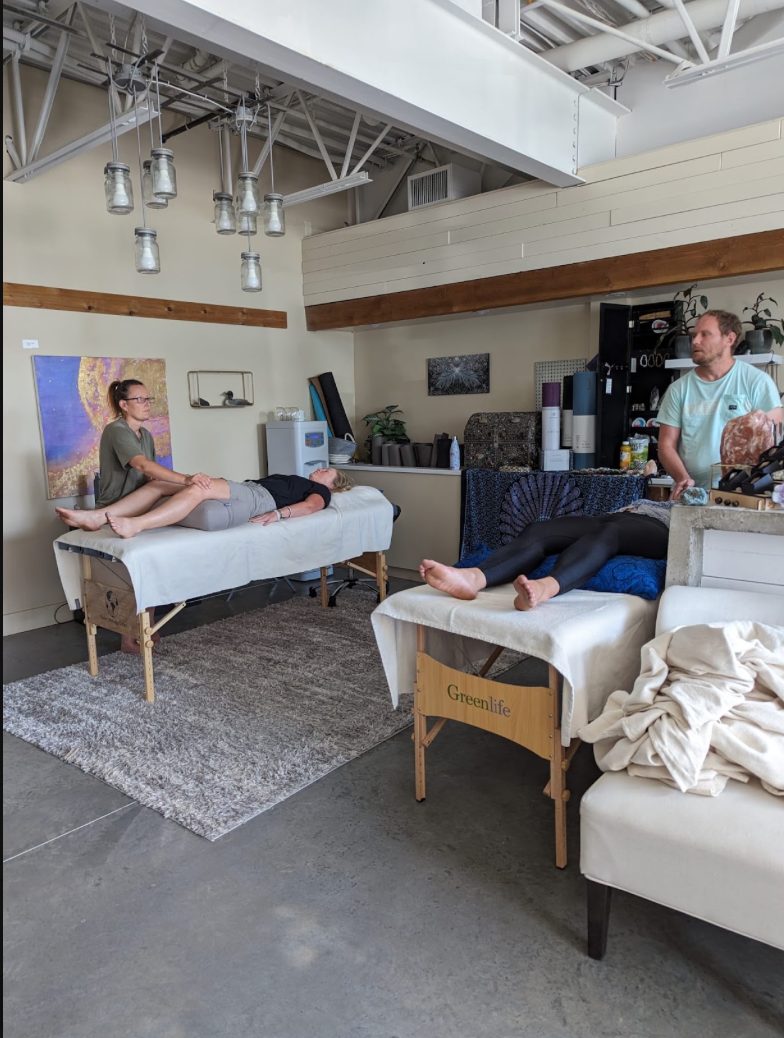 Two women, lying face-up on massage tables, receiving massages from therapists in a wellness or massage therapy studio. The studio has a modern industrial decor with hanging lights and artwork on the walls.