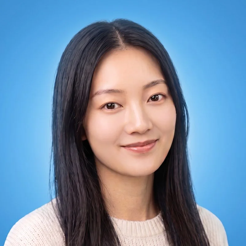 Close-up portrait of a smiling young Asian woman with long black hair, wearing a light-colored sweater, against a plain blue background.