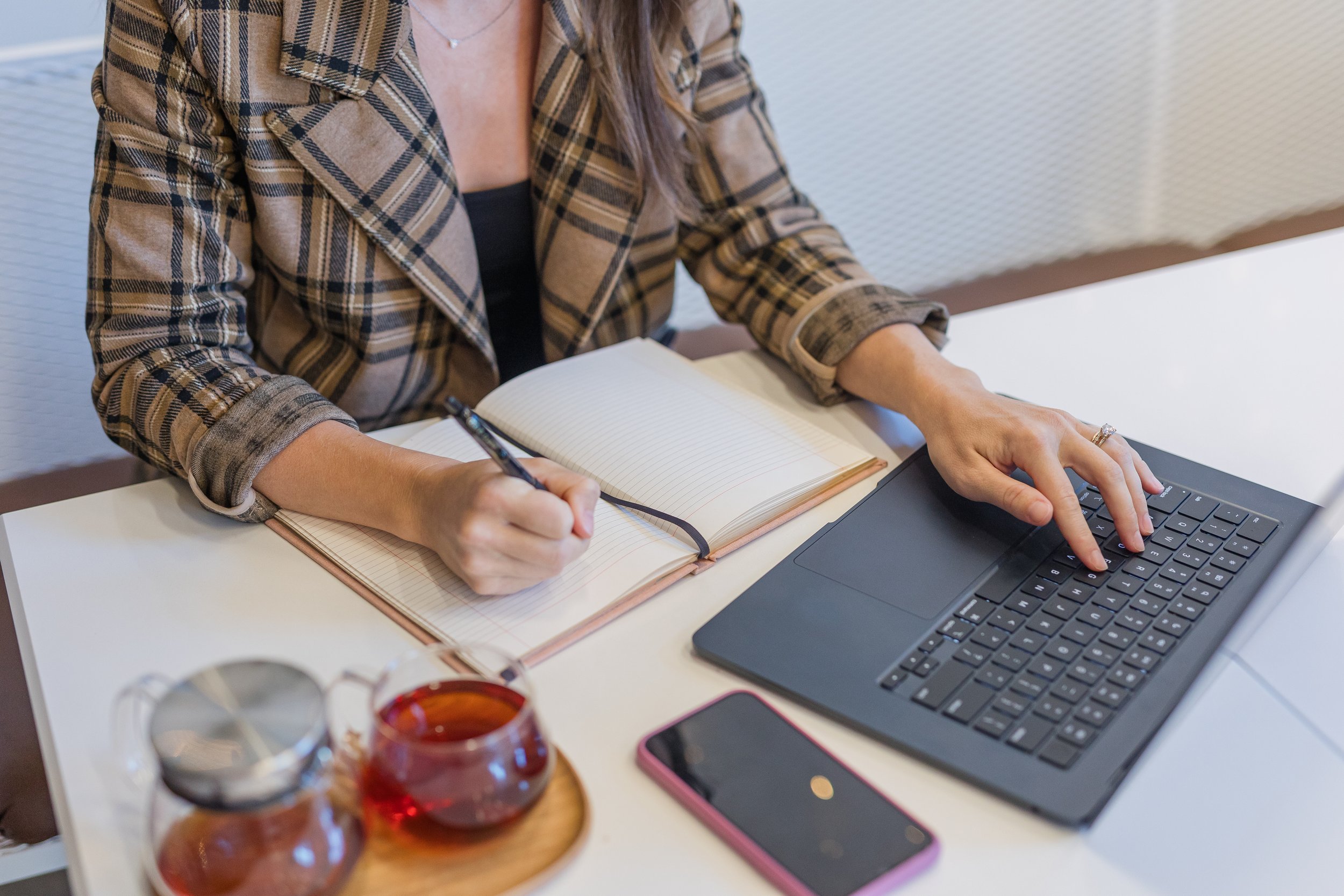 A woman sitting at a desk, writing in a notebook with a pen while using a laptop. There is a glass jar of honey or jam, and a smartphone on the desk.