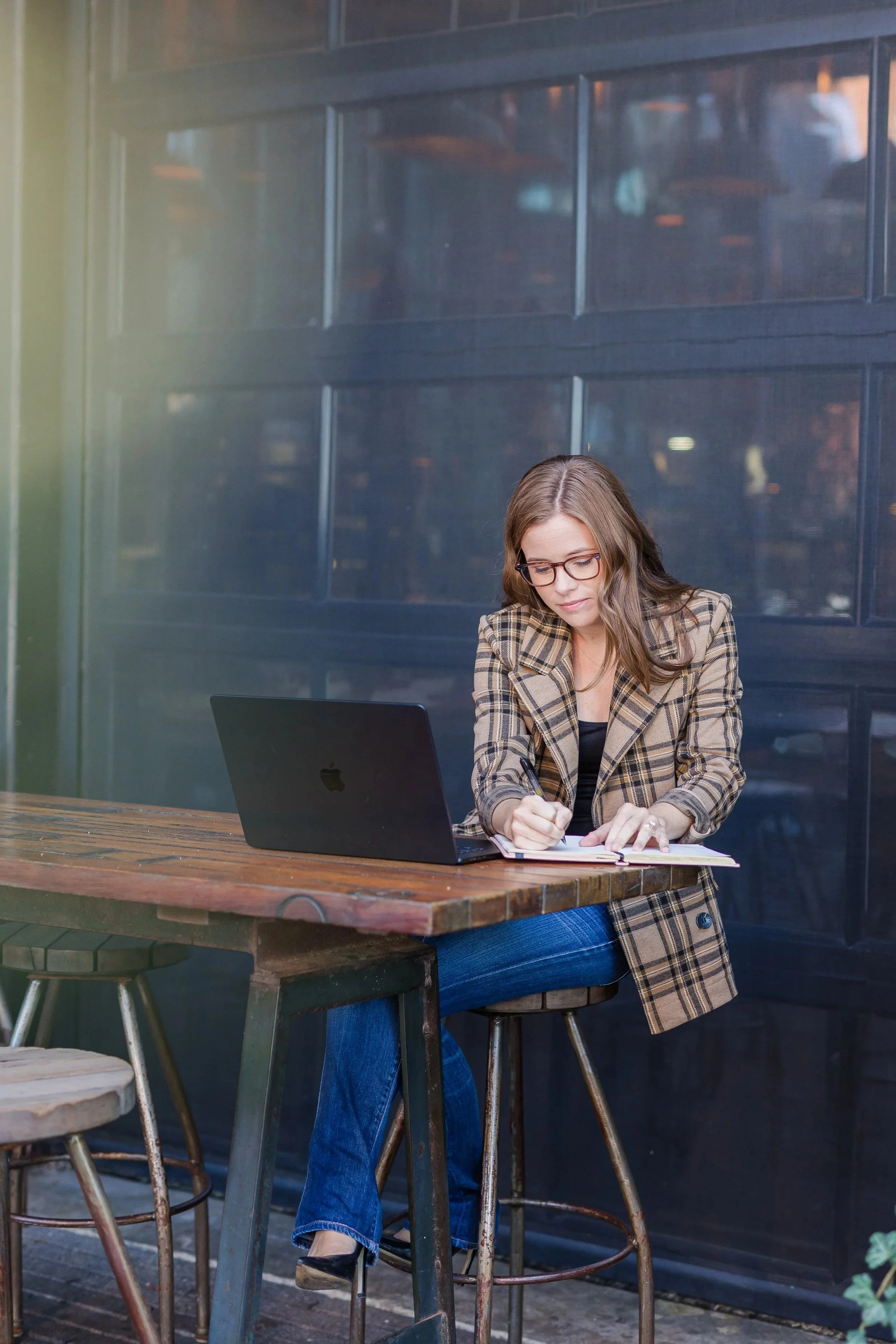 A woman with long brown hair, wearing glasses, a plaid blazer, and blue jeans, sits at a rustic wooden table outdoors, working in a notebook with a laptop nearby, in front of a glass building.