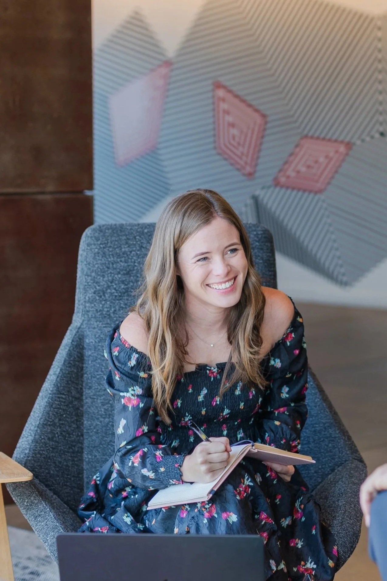 A smiling woman with wavy light brown hair sits in a gray chair, wearing a floral dress, writing notes in a notebook.