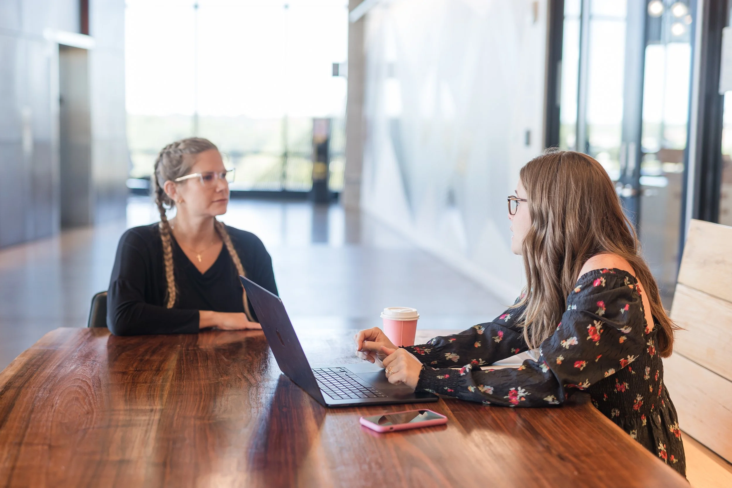 Two women sitting at a wooden table in a modern, brightly lit coffee shop, engaged in conversation with laptops and a coffee cup on the table.