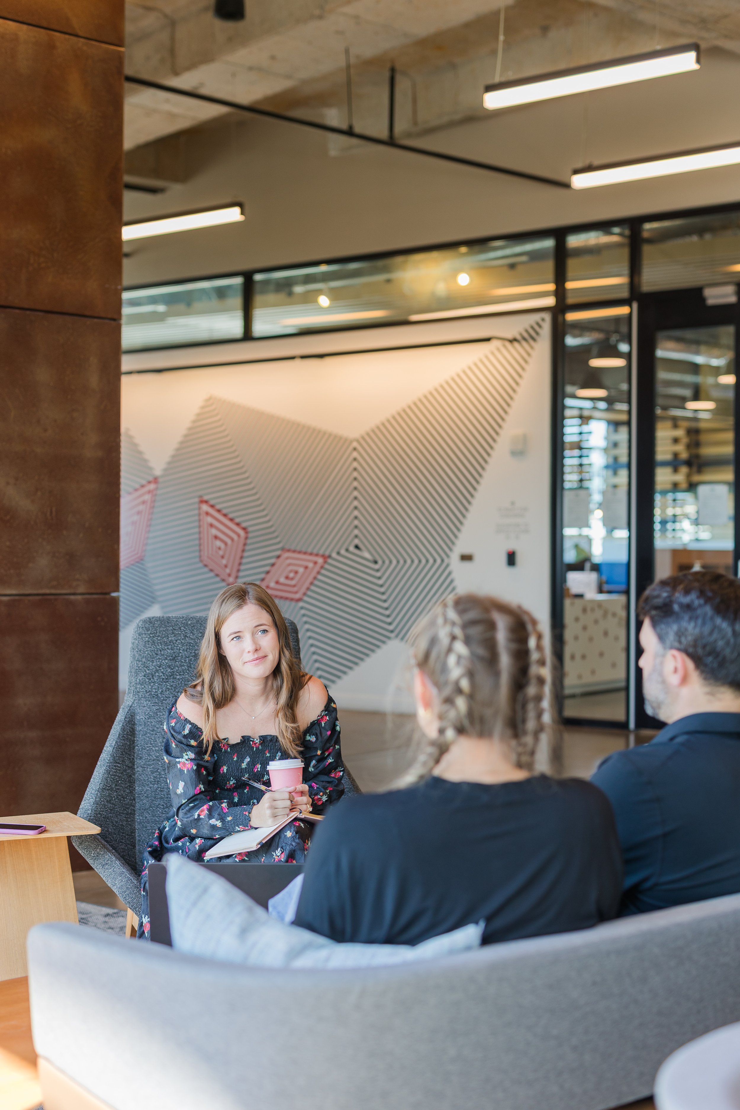 A group of young adults, twomen and one man, sit around a table in a modern office or coworking space. The woman in floral dress is holding a pink coffee cup, and all are engaged in conversation. The background shows a large wall with geometric art and glass walls with metal framing.