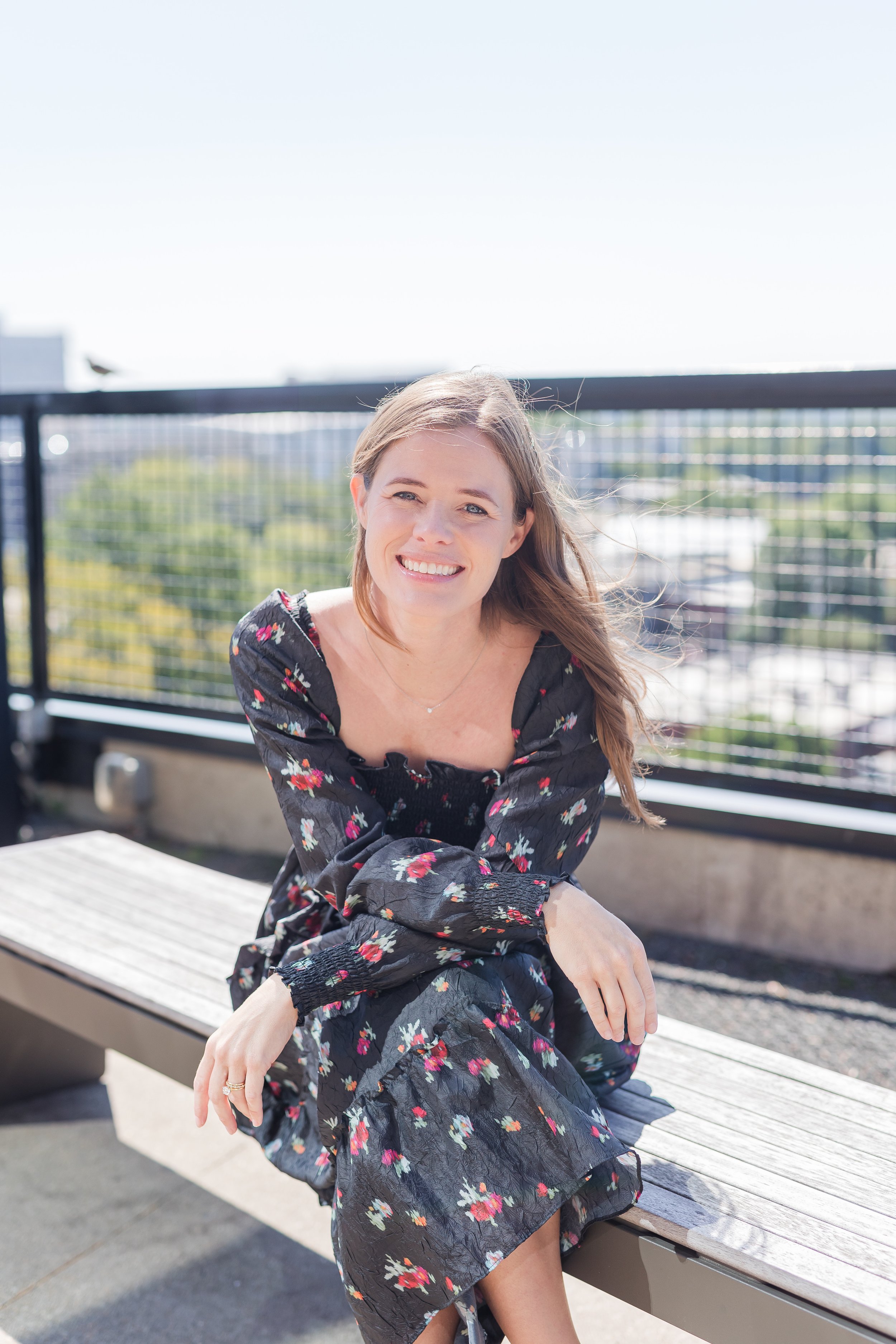 A smiling woman with long, light brown hair in a floral black dress with long sleeves, sitting on a wooden bench outdoors on a sunny day, with a black metal railing and blurred cityscape in the background.