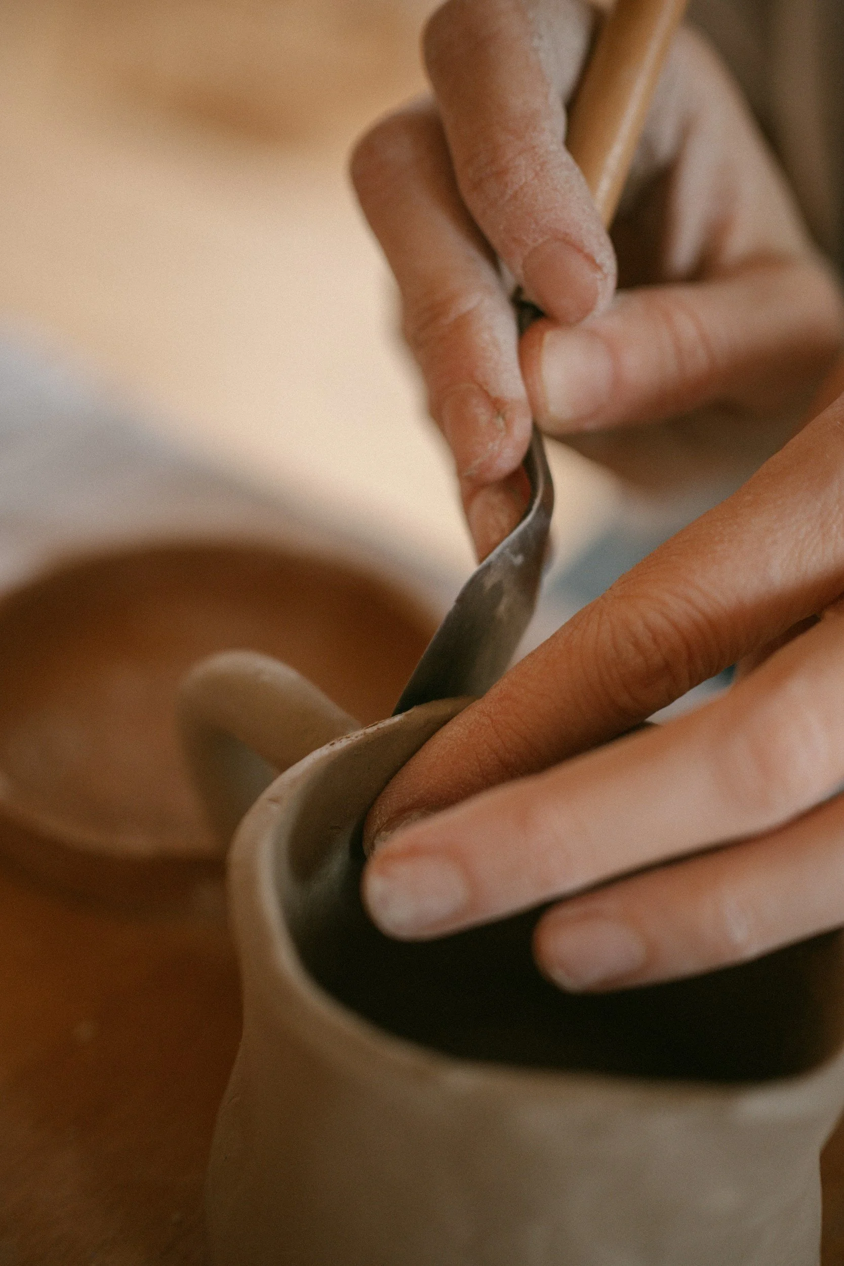 Close-up of a person's hand carving a design into a ceramic mug with a small carving tool.