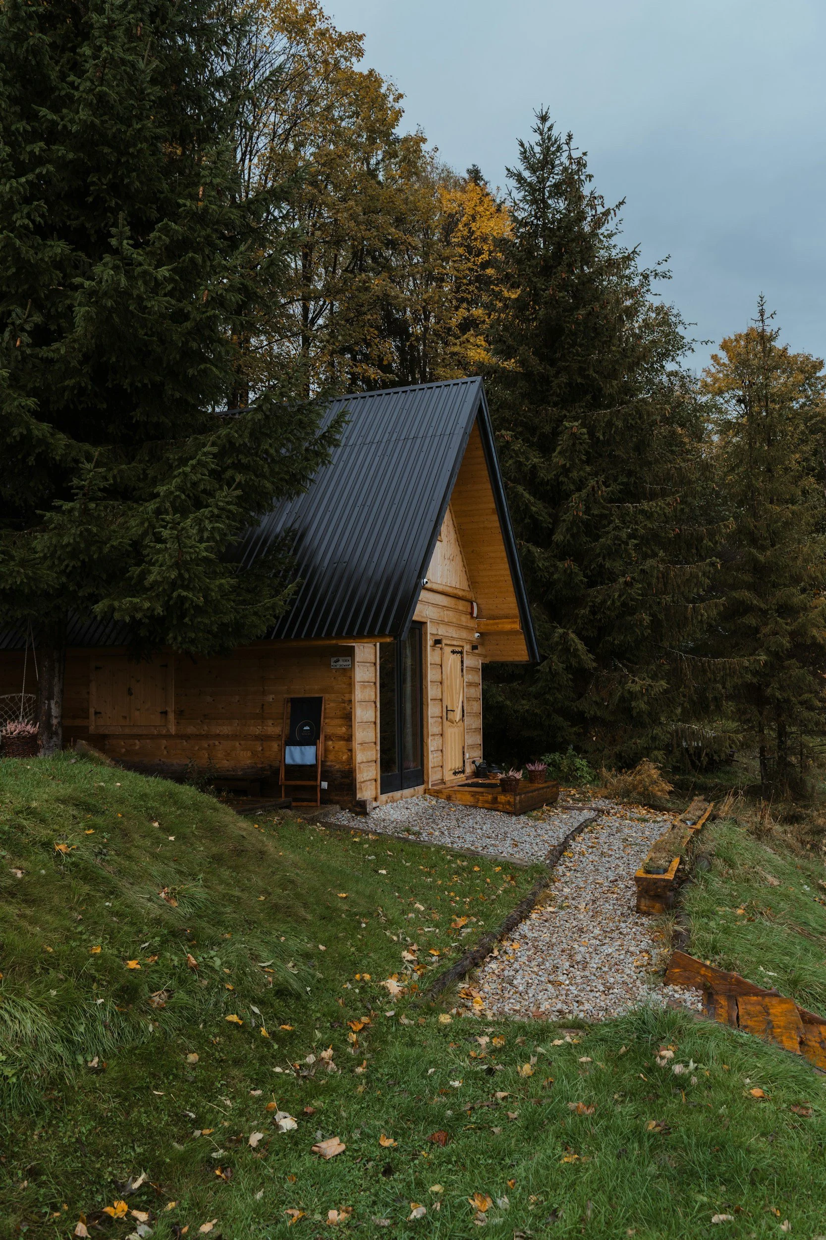 A wooden house with a steep black metal roof surrounded by tall pine trees and some deciduous trees with yellow leaves, on a partly cloudy day.