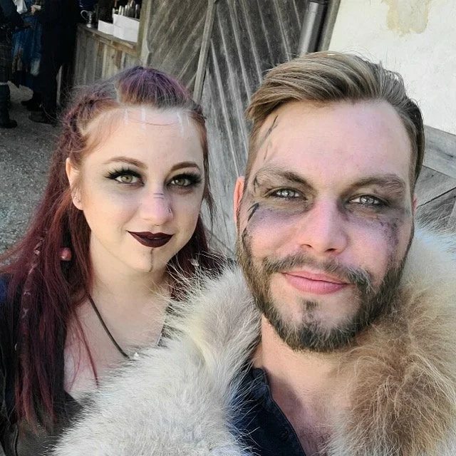A man and woman in costume with makeup, posing for a selfie outdoors. The woman has dark lipstick, eye makeup, and purple hair. The man has makeup around his eyes and a furry coat.