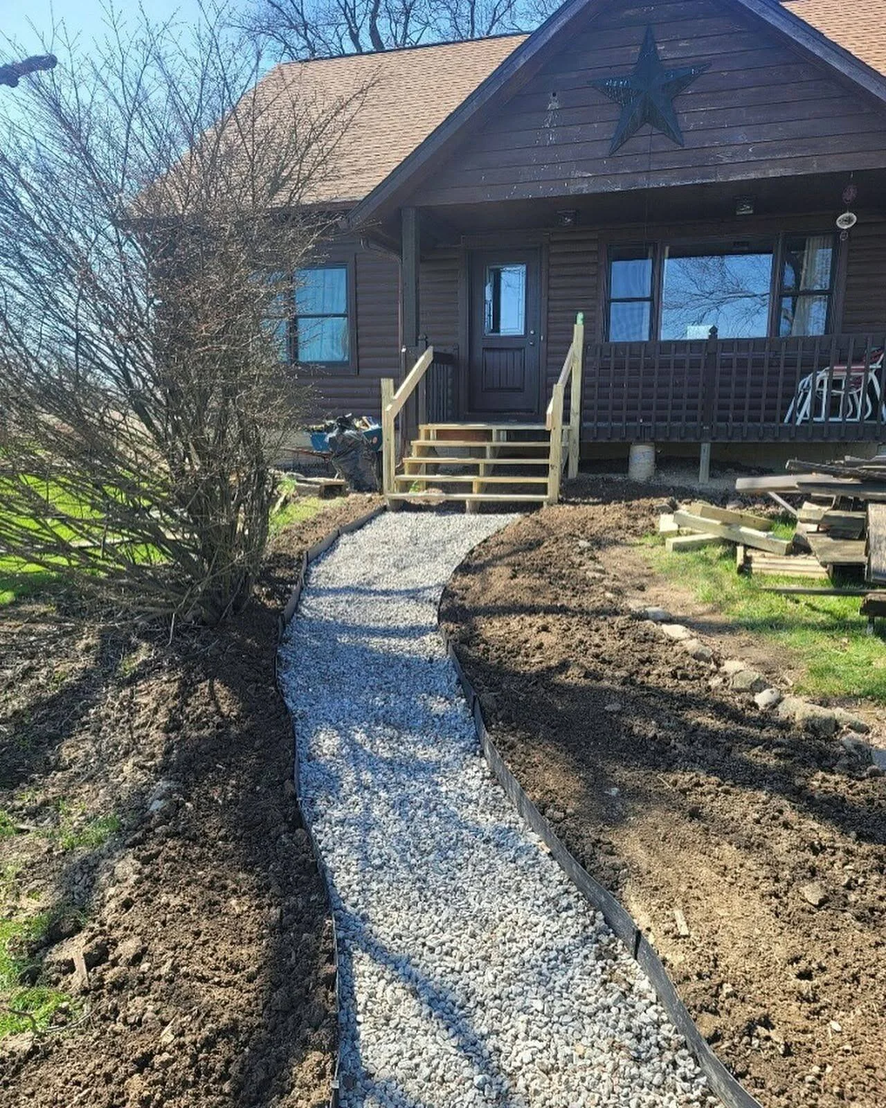 A newly laid gravel pathway leading up to a wooden porch with stairs on a house. The house is brown with a large star decoration on the gable end. There are trees and landscaping work in progress around the pathway.