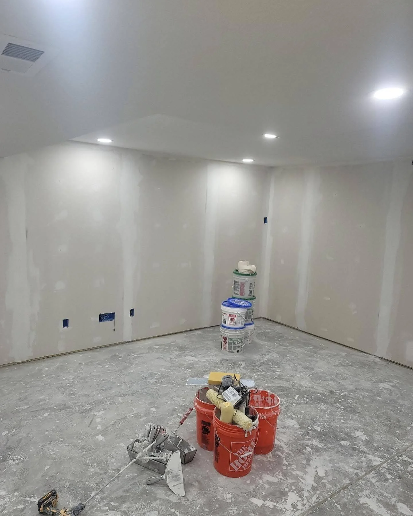 Room under construction with drywall on walls, ceiling lights installed, buckets of drywall compound or paint, and construction tools and materials on the unfinished floor.
