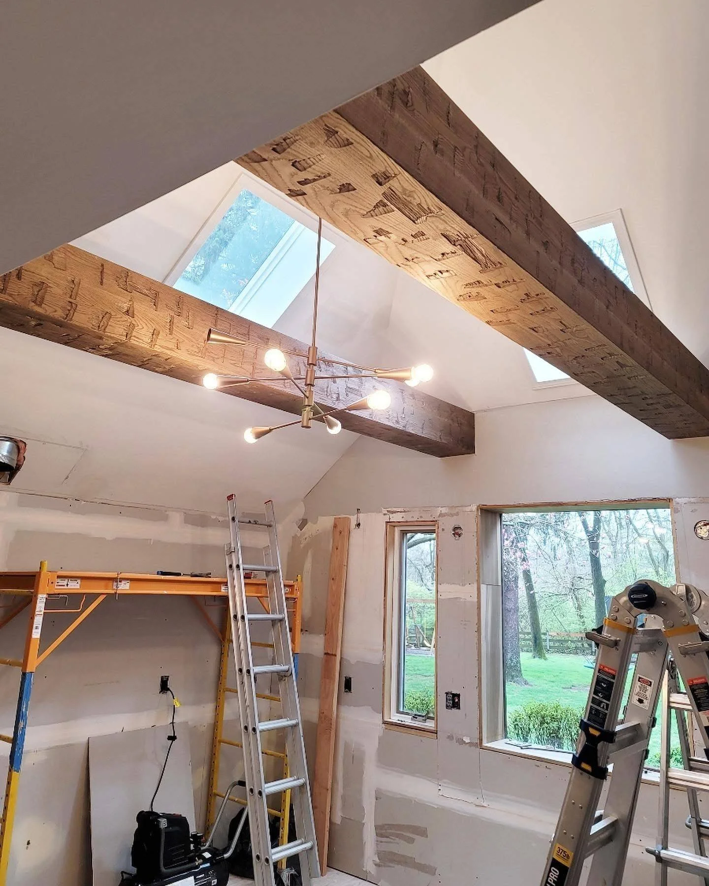 Interior of a house under renovation with exposed beams, construction tools, ladders, and large windows showing a view of trees outside.