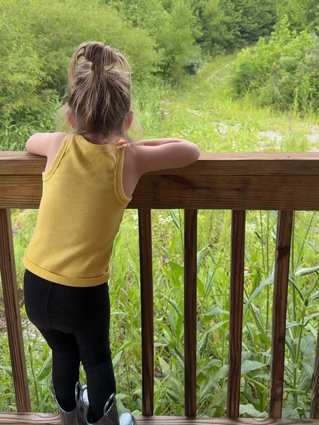 Child with a yellow sleeveless top and black pants leaning on wooden railing, looking into green forested area.