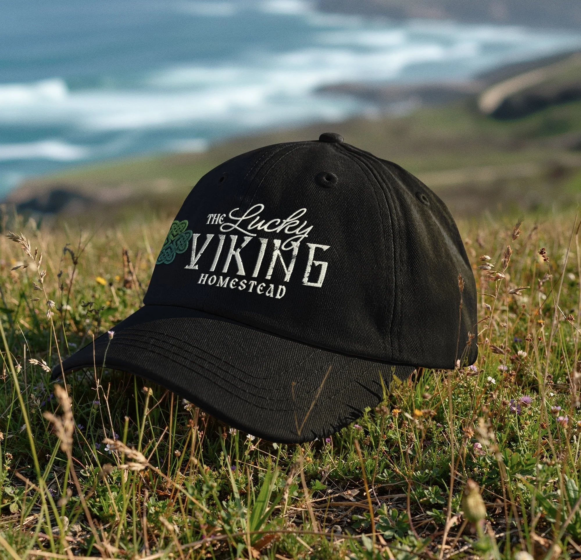 A black baseball cap with white embroidered text reading "The Lucky Viking Homestead" rests on grass with small wildflowers; in the background, the ocean with blue water and waves is visible under a cloudy sky.