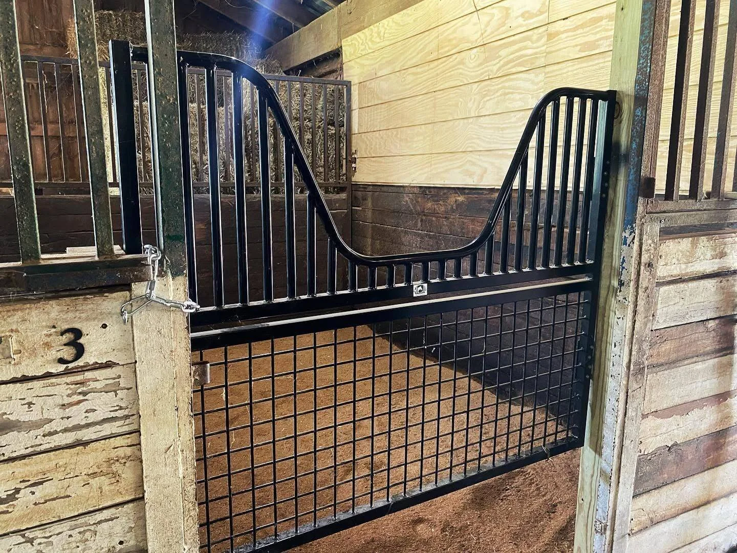 A black metal horse stall door with a latch, installed in a wooden stable wall with light and dark wood panels.
