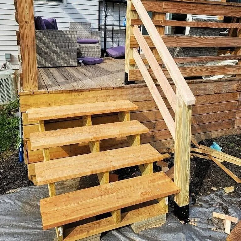 Newly built wooden staircase leading to an outdoor deck with patio furniture. The steps are unfinished, with shadows indicating sunlight.
