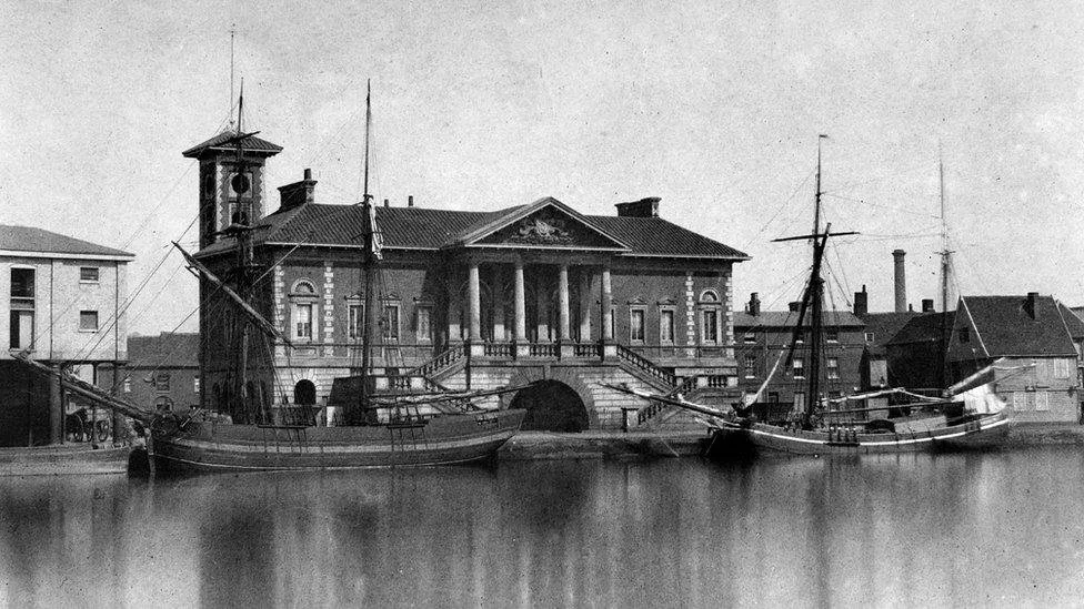 Black and white photograph of two boats docked in front of a historic building with columns and stairs leading to the entrance.