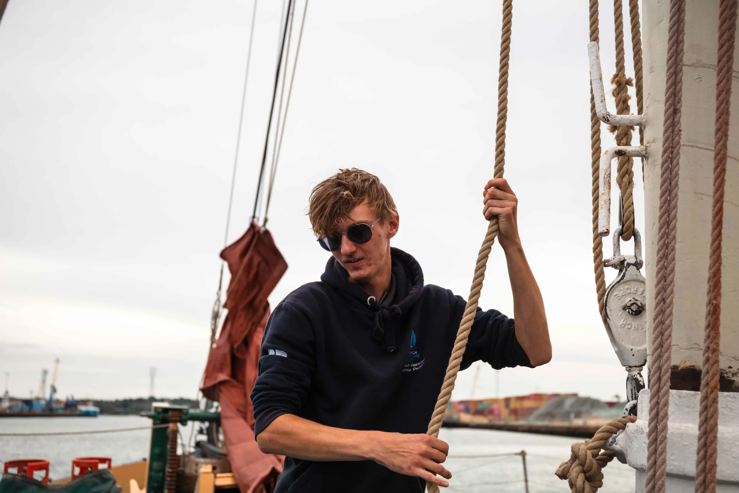 A young man with glasses and brown hair, smiling, holding onto a rope on a boat during sunset, with other boats and water in the background.