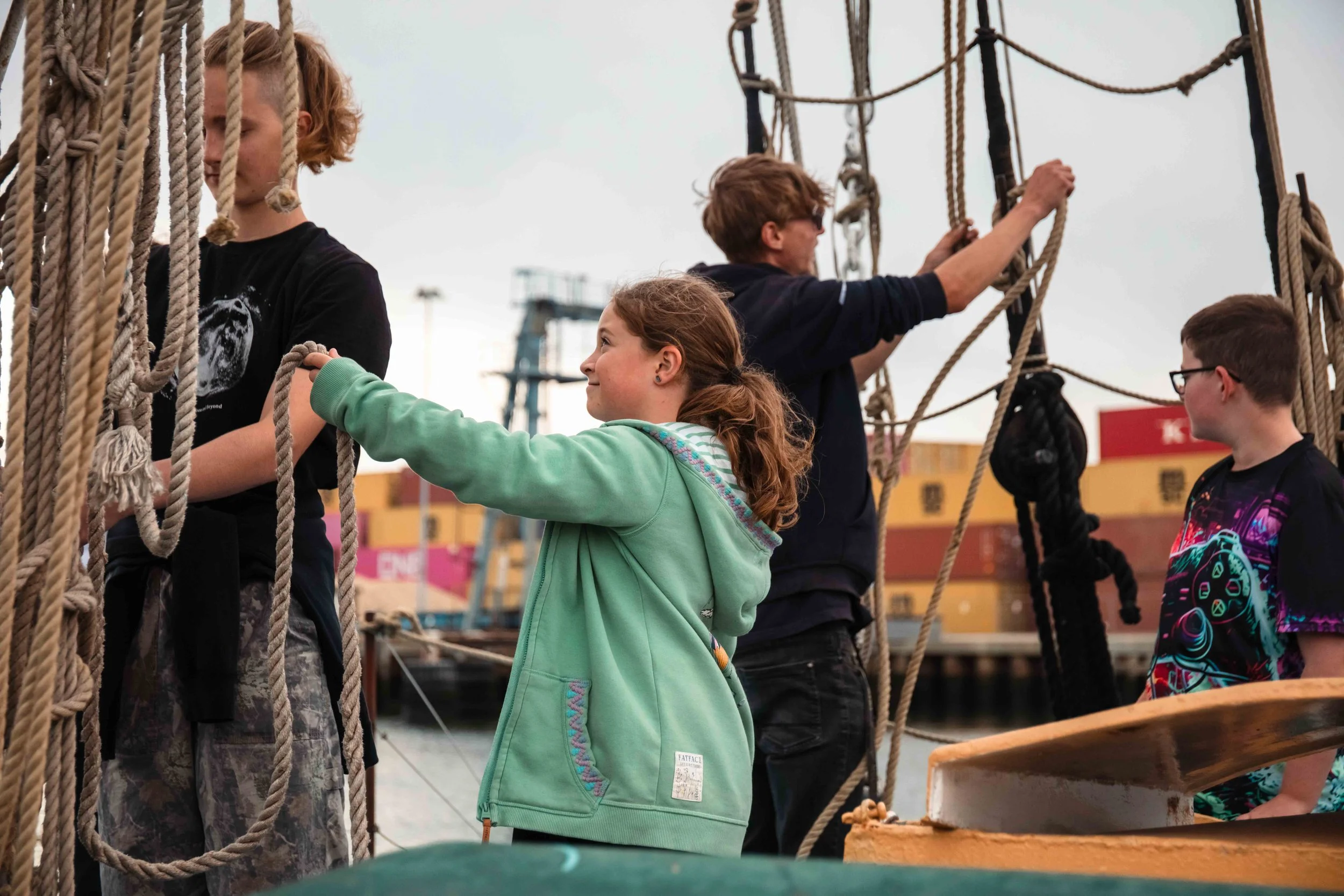 Children and adults on a ship's rope structure, with some holding ropes, at a harbor with boats and industrial buildings in the background.