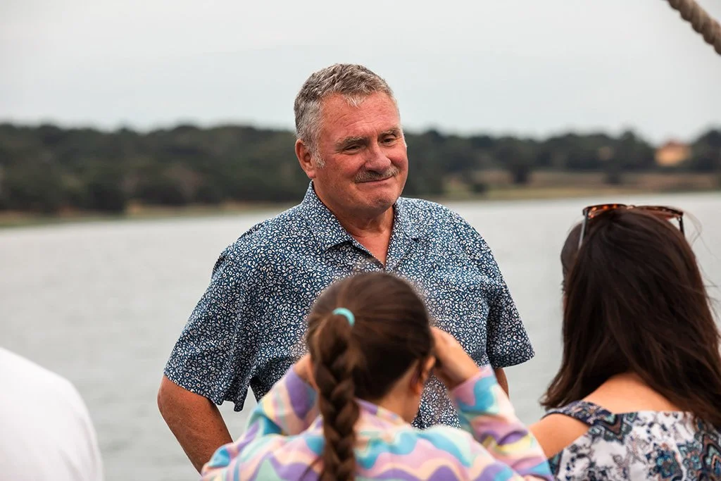 A man with gray hair in a patterned short-sleeve shirt smiling, standing outdoors near a body of water, talking to two women and a girl with braided hair.