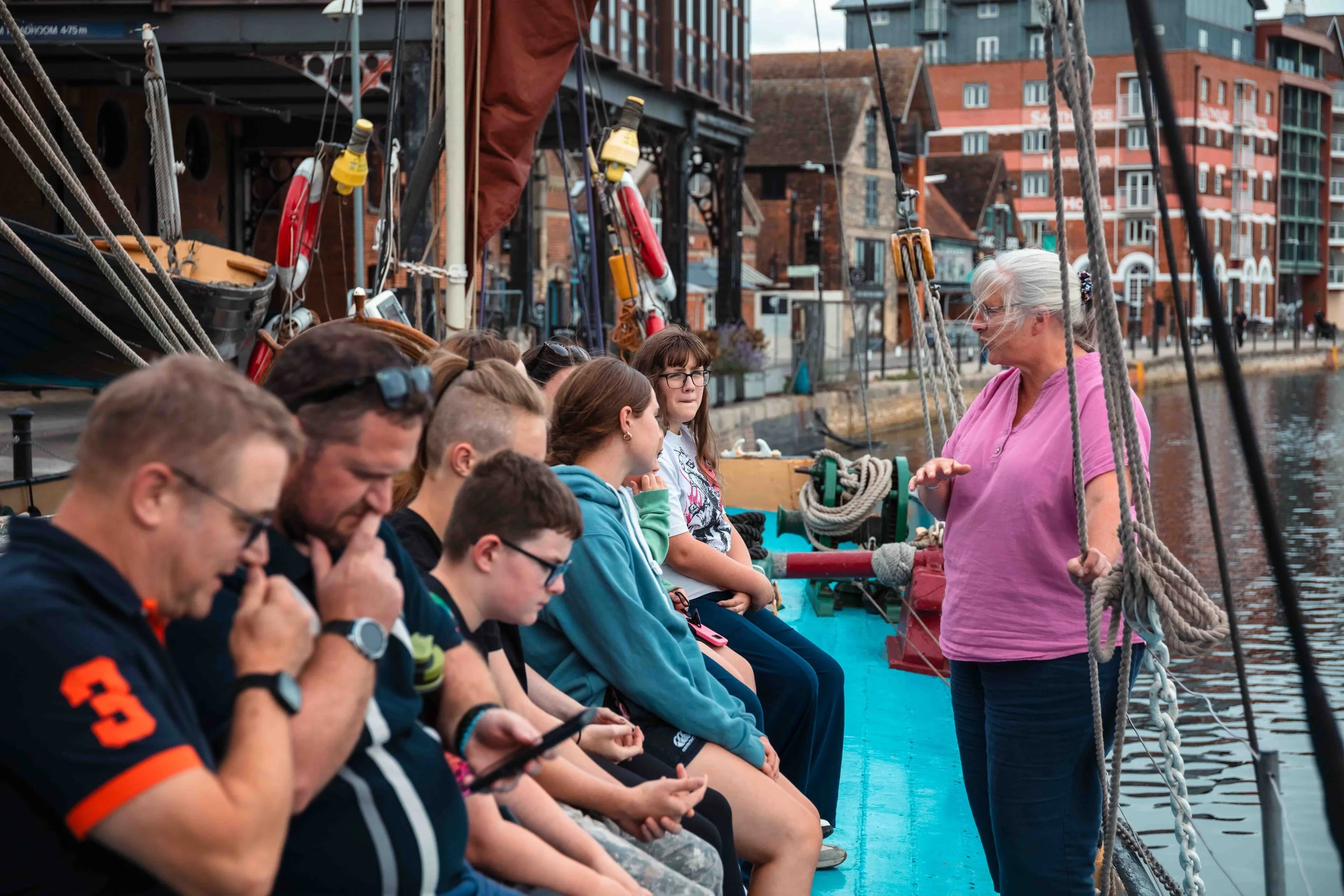 People sitting and standing on a boat docked at a harbor, with some reading and talking, and a woman walking nearby.