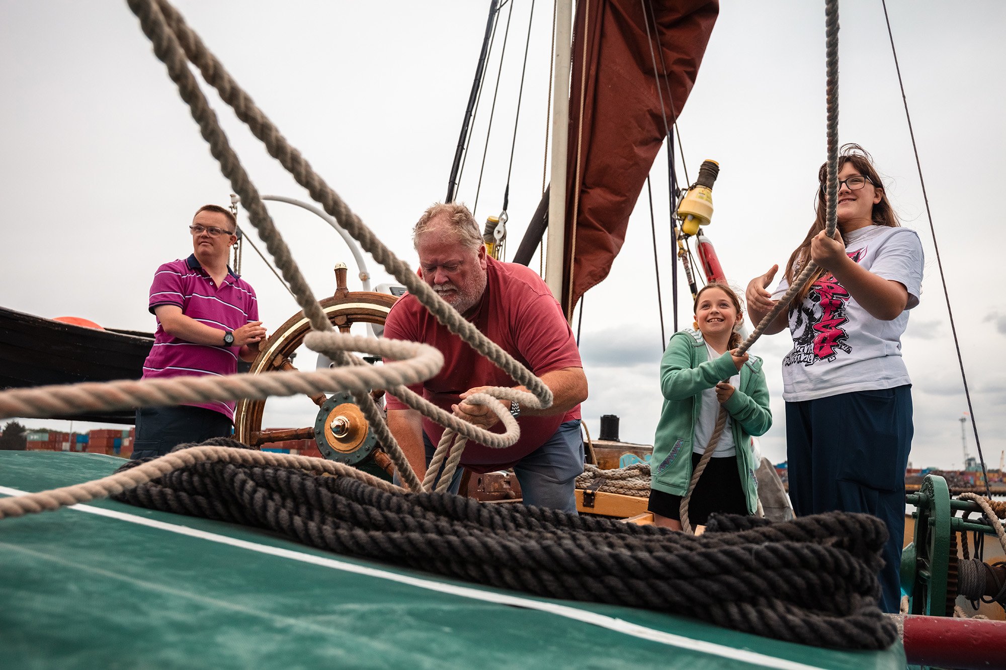 A group of people on a docked sailboat, with some sitting and others standing, engaged in activities like reading or using devices, in a harbor setting with buildings and other boats in the background.