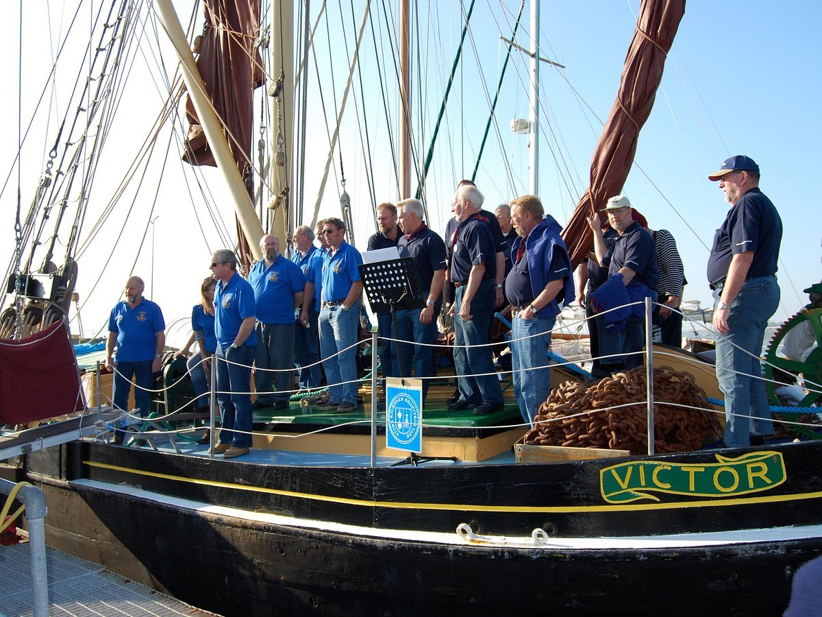 Group of people standing on a black boat named Victor, with ropes and rigging, in a marina during the day.